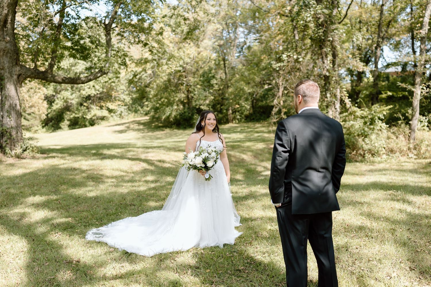 A bride in a flowing white dress holds a bouquet while standing with a person in a black suit in a sunlit garden setting.