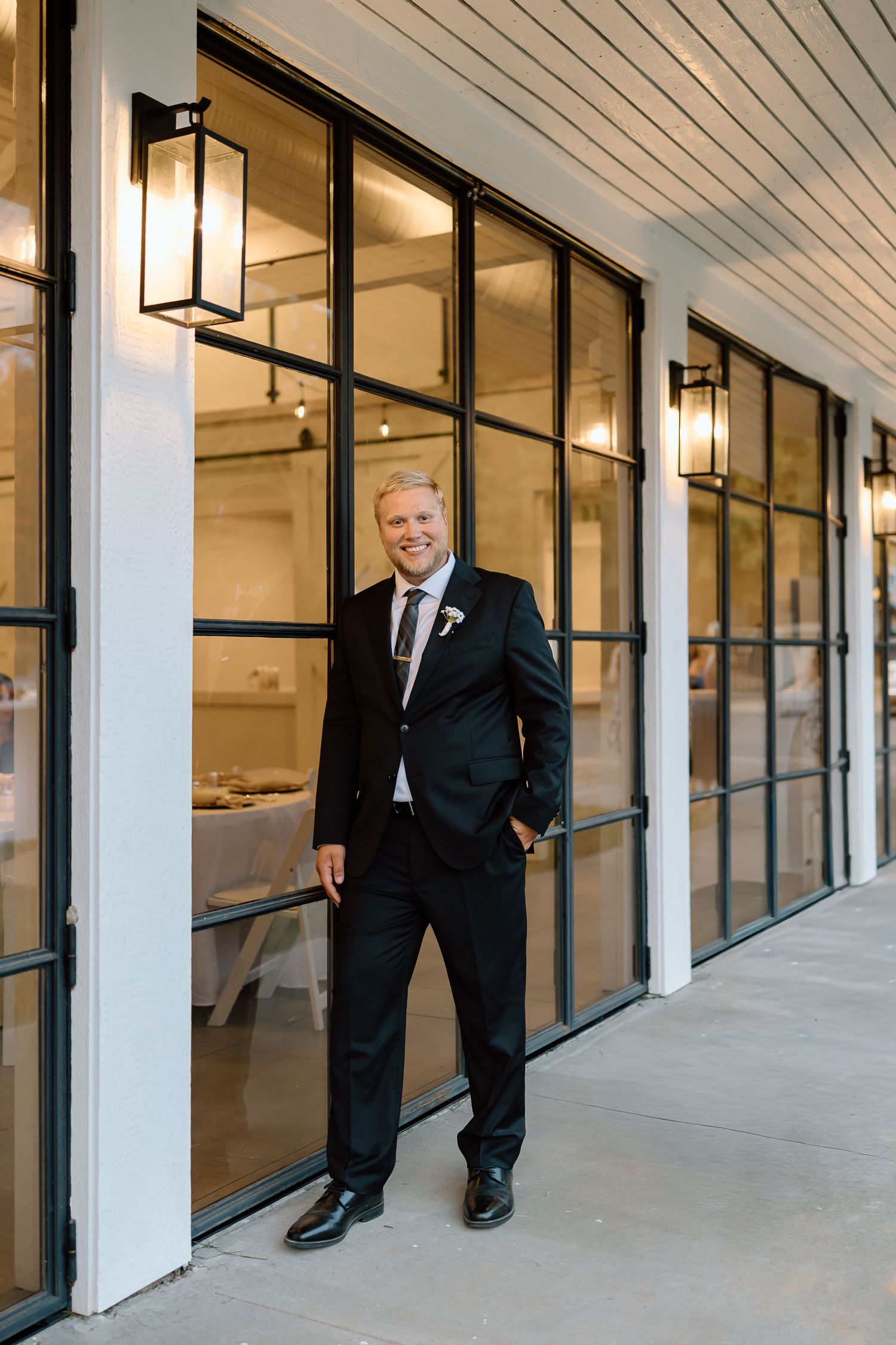 A person in a black suit stands in a modern office hallway with white columns and glass walls.
