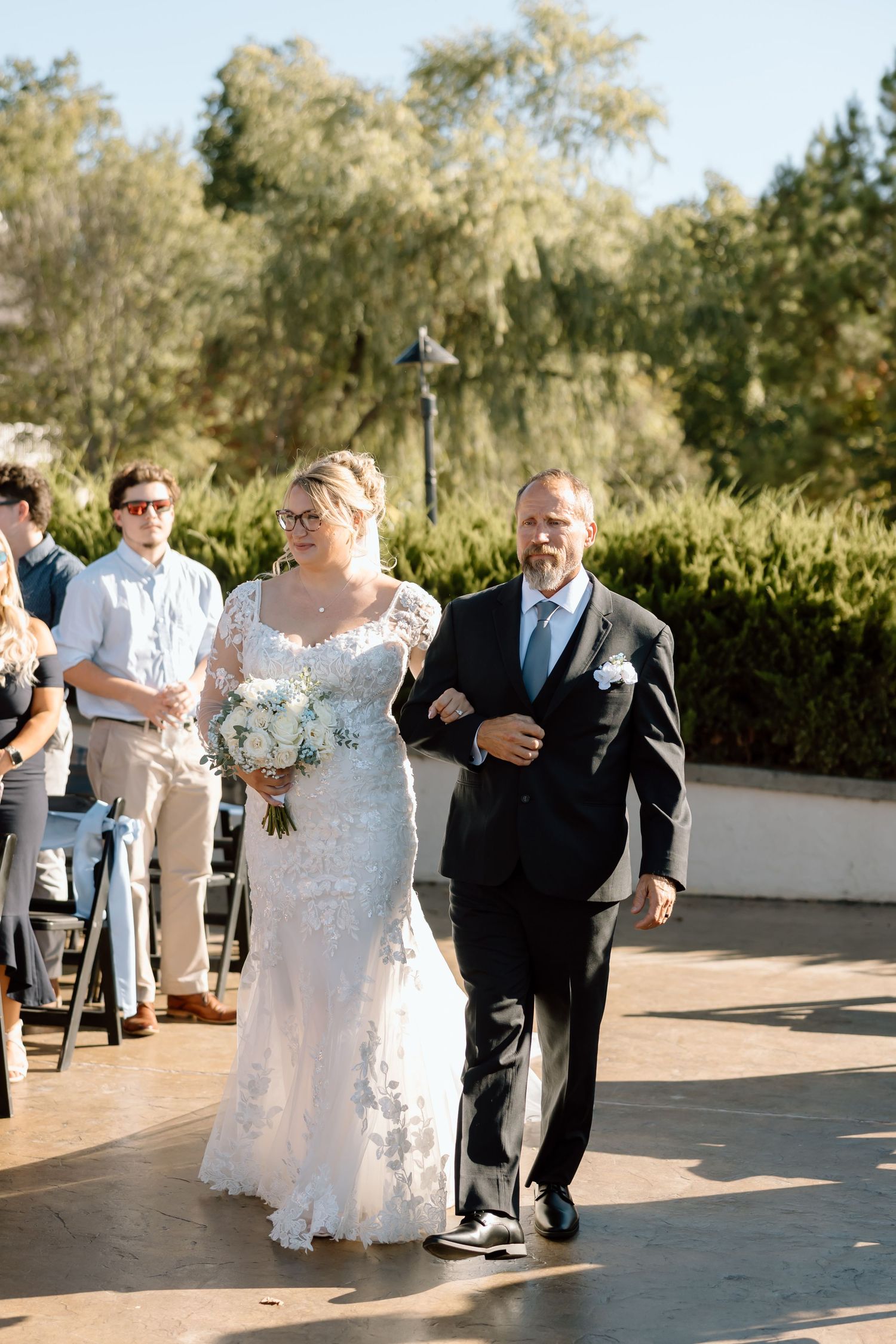 A couple dances together during an outdoor wedding reception on a sunny patio surrounded by greenery.