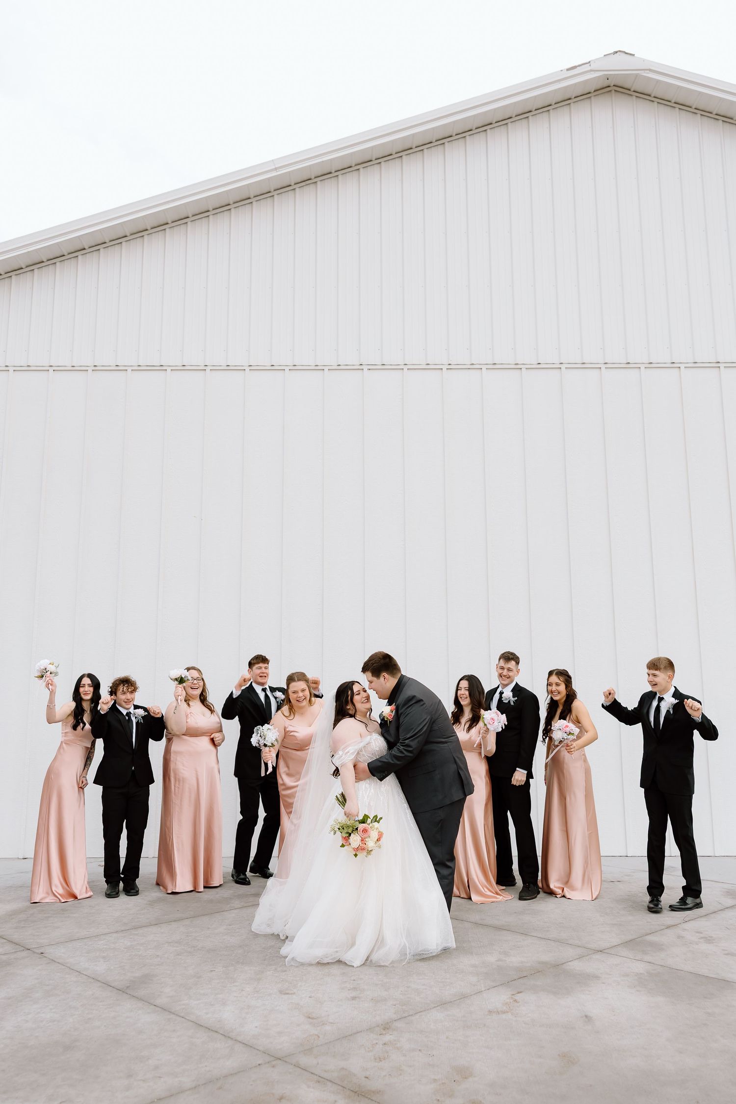 Wedding party in pink bridesmaid dresses and black suits poses playfully against white wall in creative lineup sequence.