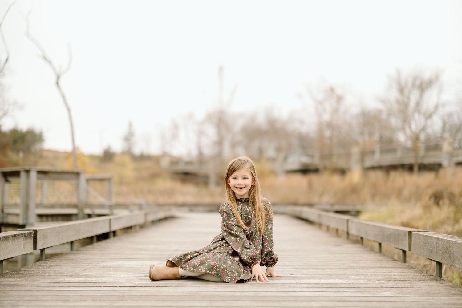 A child in a floral dress sits gracefully on a wooden boardwalk during a photo session.