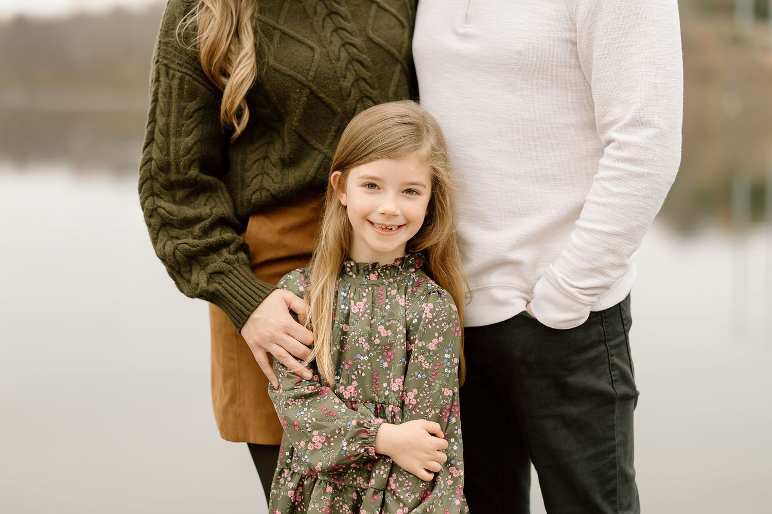 Young girl in floral dress stands with parents on wooden dock by serene lake during autumn.