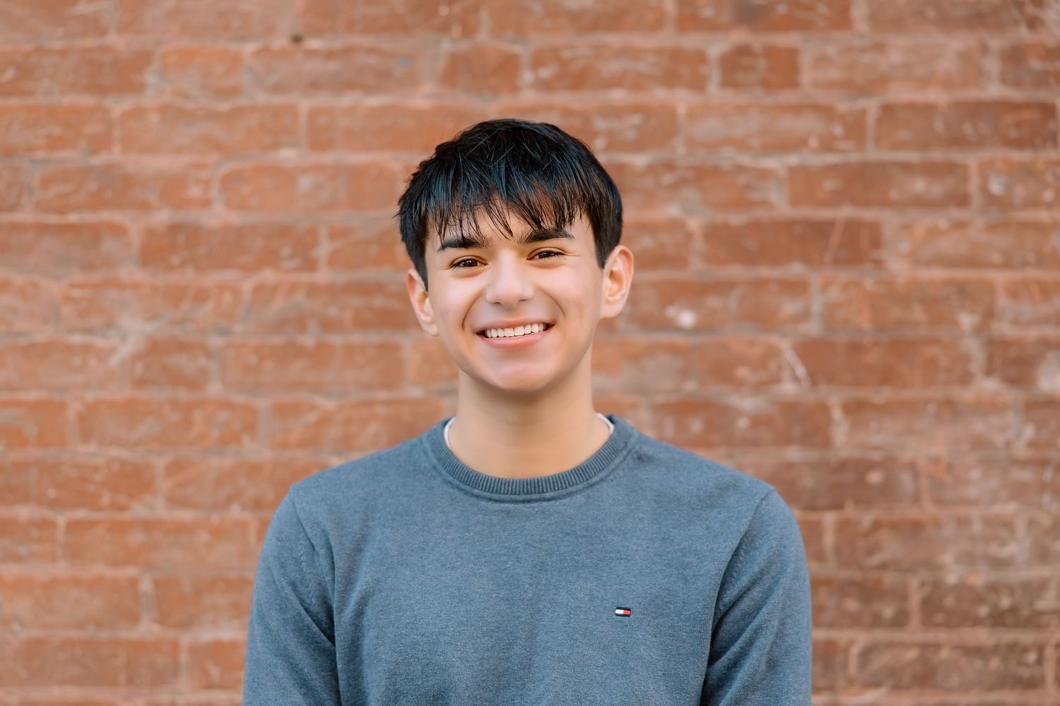 Close-up portrait in front of red brick wall wearing a casual blue Tommy Hilfiger sweater.