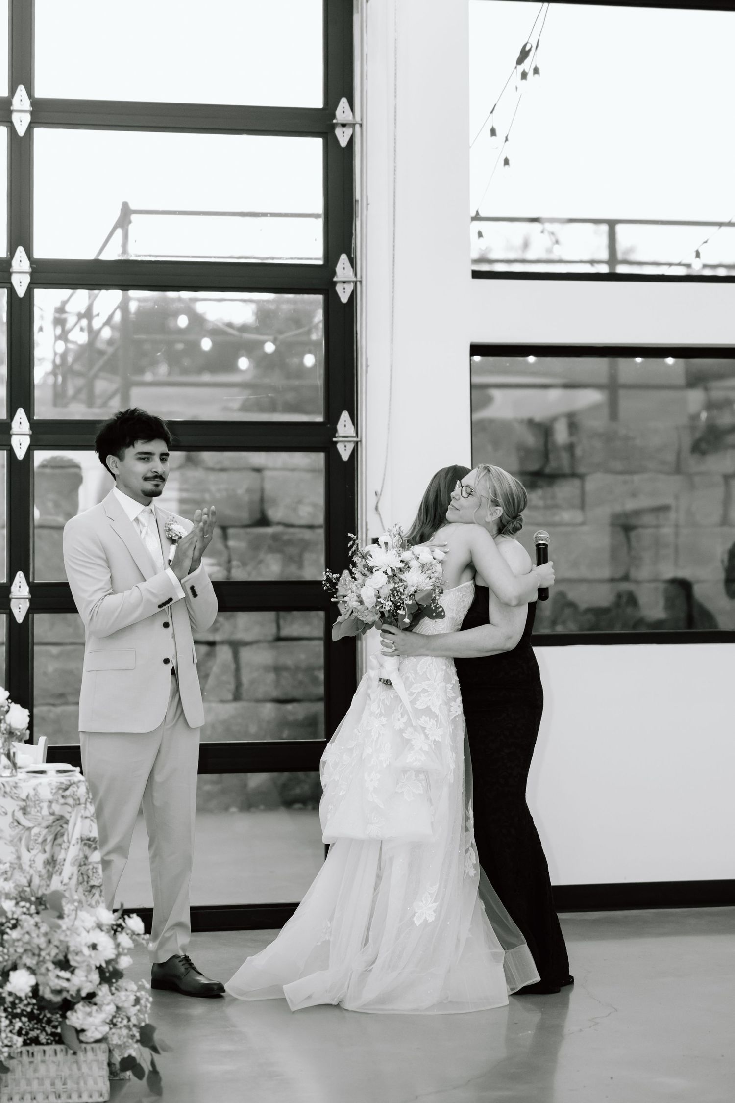 Bride hugging a guest while groom watches during wedding reception in elegant venue with large windows.