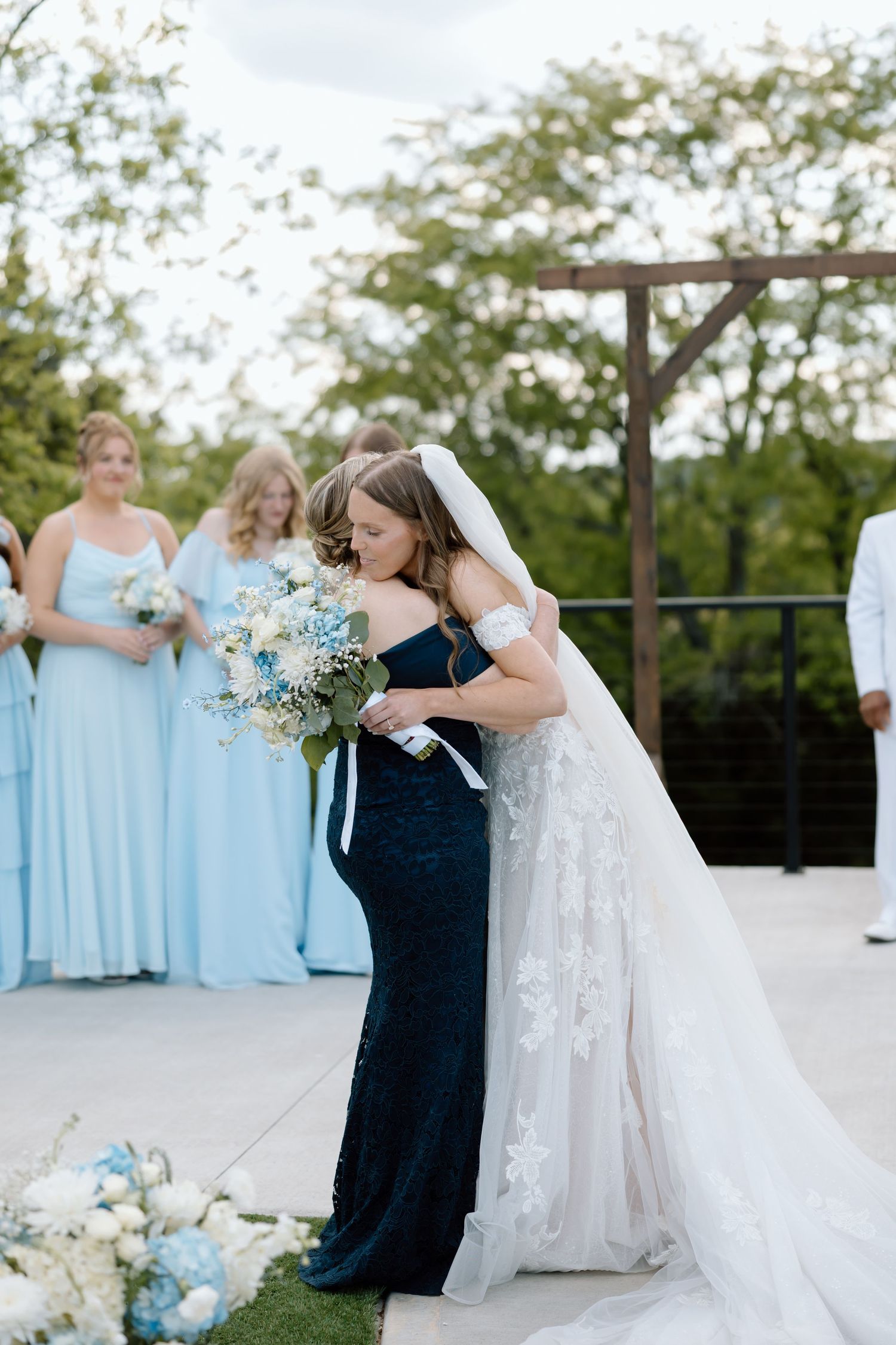 Bride in lace gown hugging bridesmaid in navy dress at outdoor wedding ceremony with light blue bridesmaids in background.