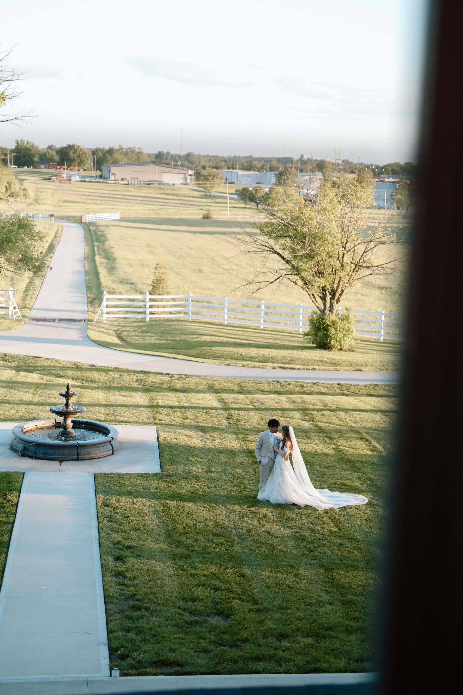 Aerial view of a bride and groom embracing on lush green lawn near a fountain at an elegant countryside wedding venue.