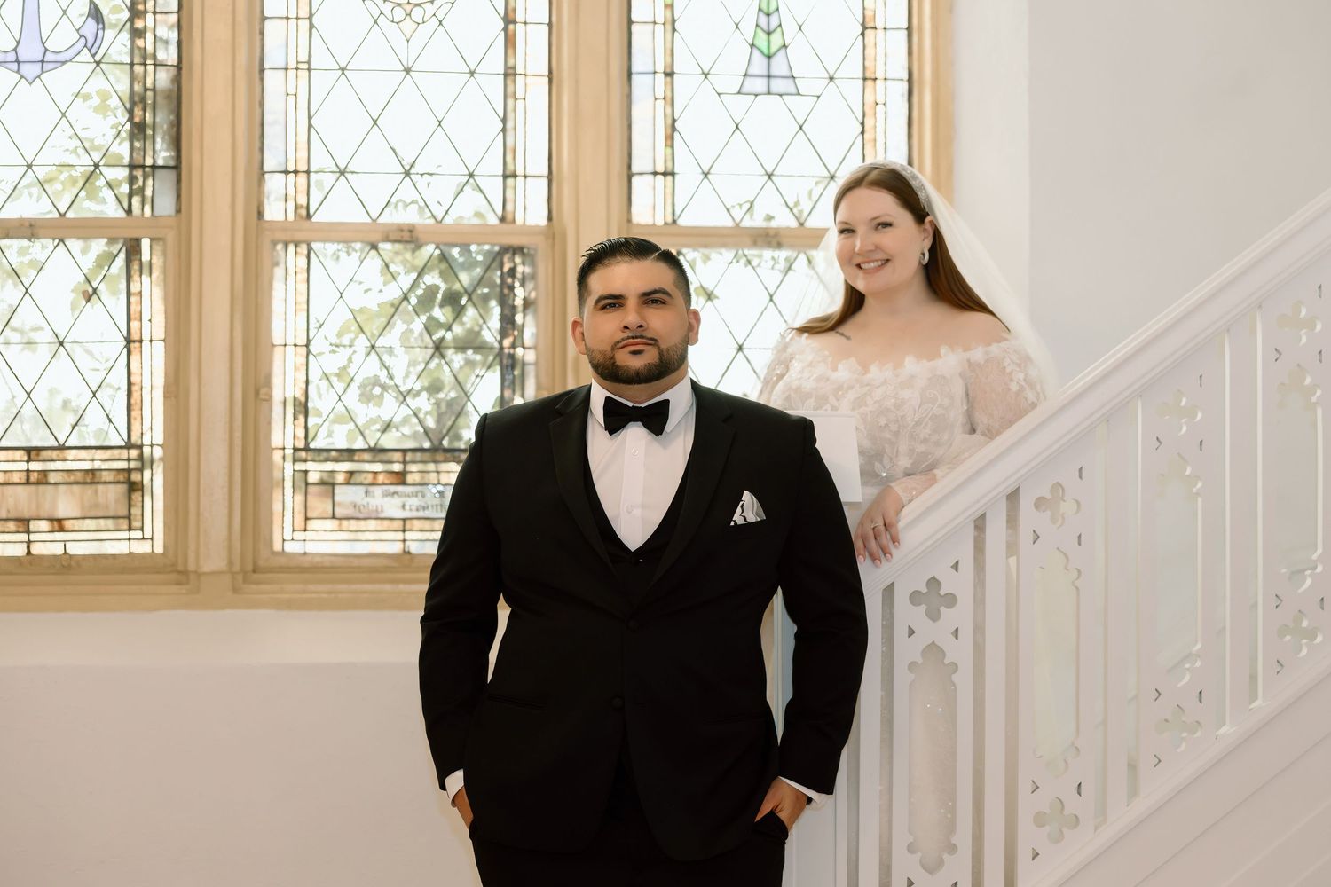 Bride in white lace gown stands on staircase behind groom in black tuxedo near stained glass windows.