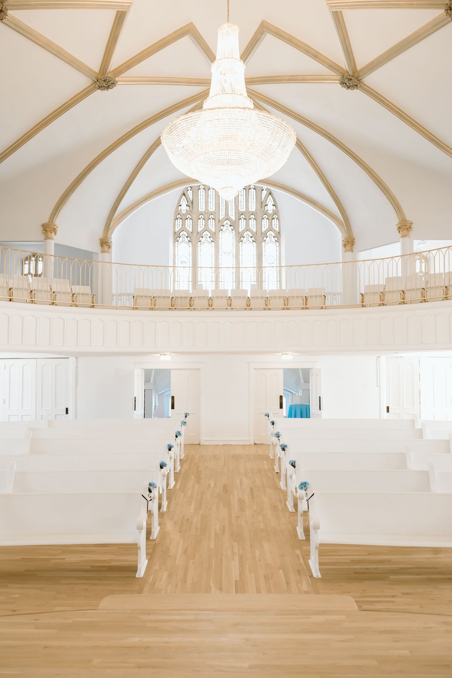 Elegant white chapel interior with vaulted ceilings, crystal chandelier, wooden pews, and Gothic stained glass window.