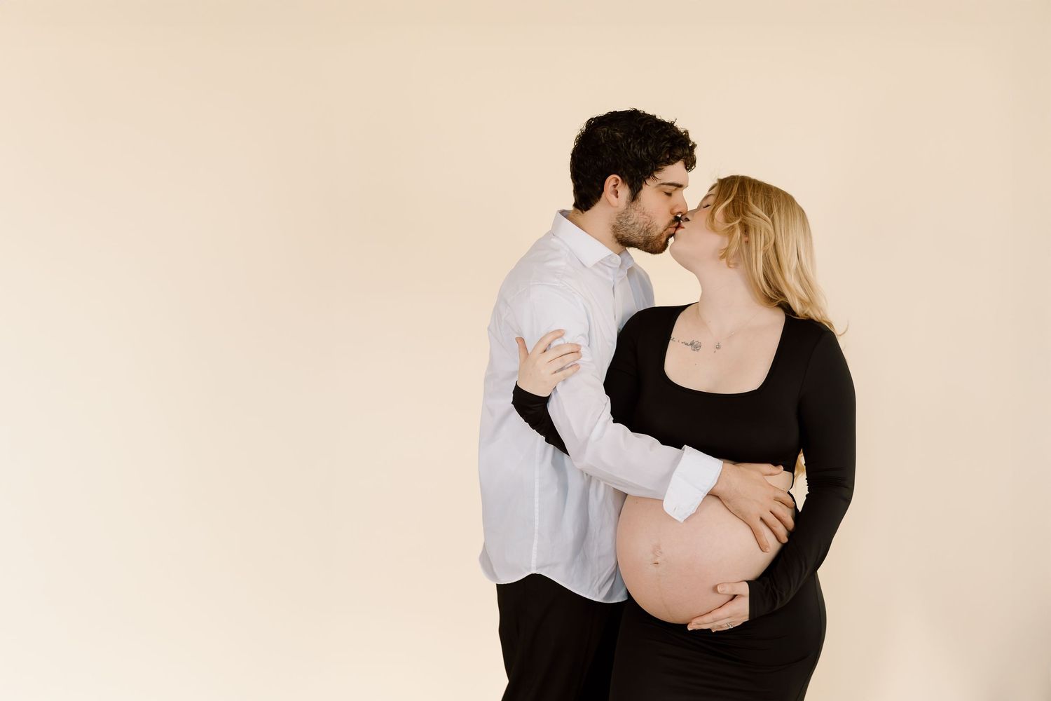 A couple embraces tenderly against a beige backdrop during a maternity photoshoot.