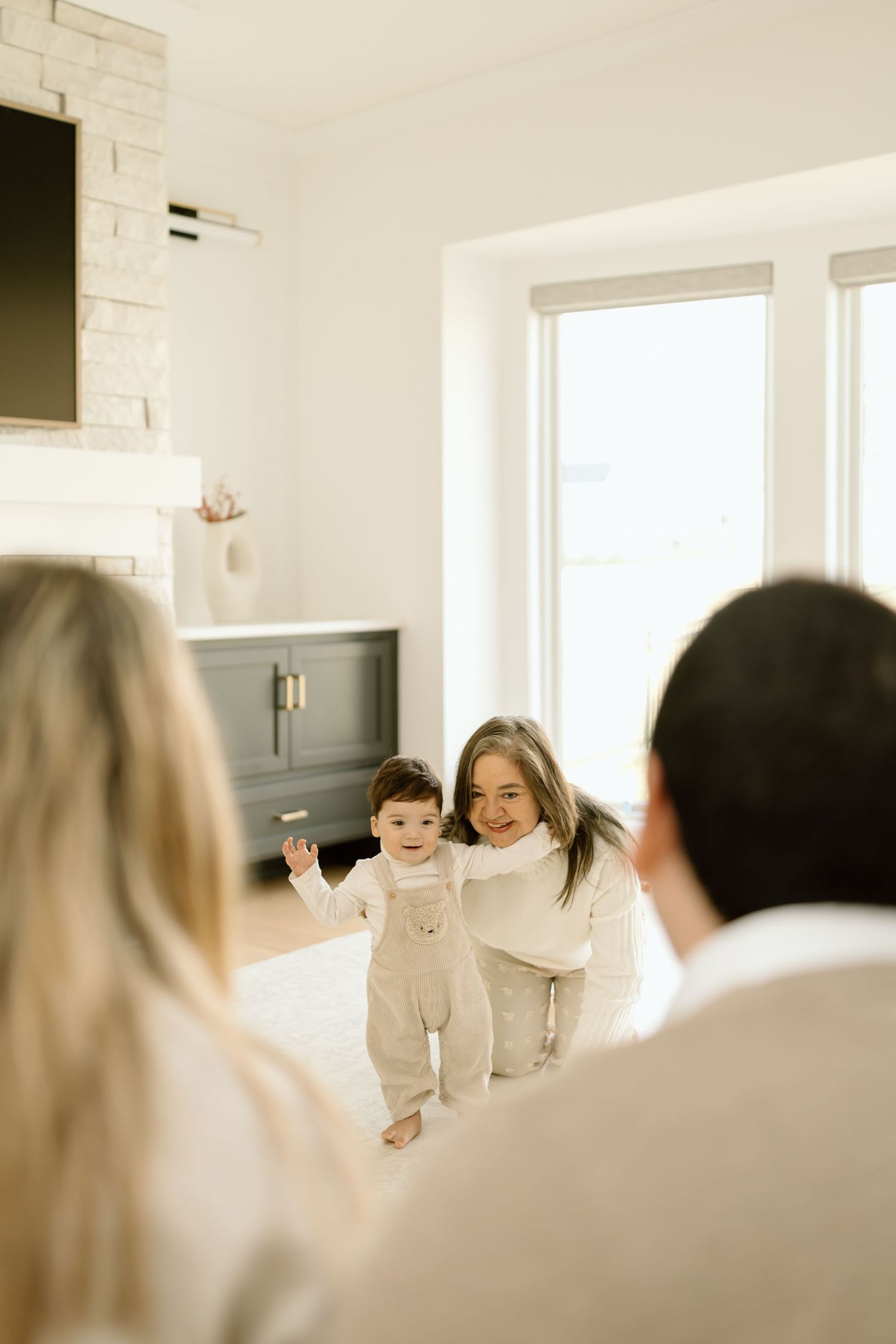 A baby in white crawls across a bright sunlit room with hardwood floors and white walls.