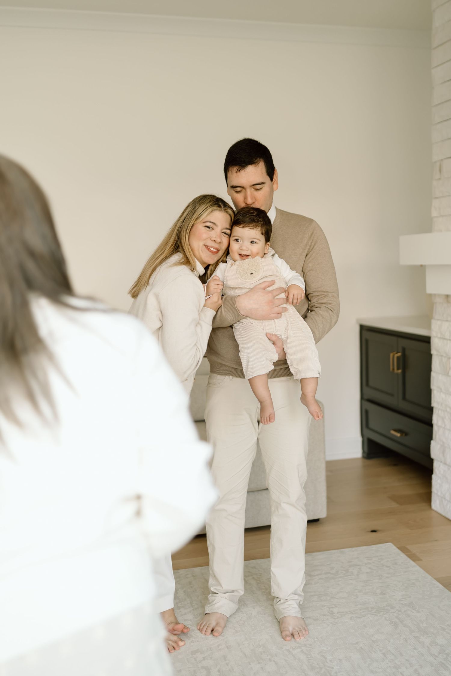 Family wearing neutral colored clothing cuddles together on a light carpet in a bright minimalist room.