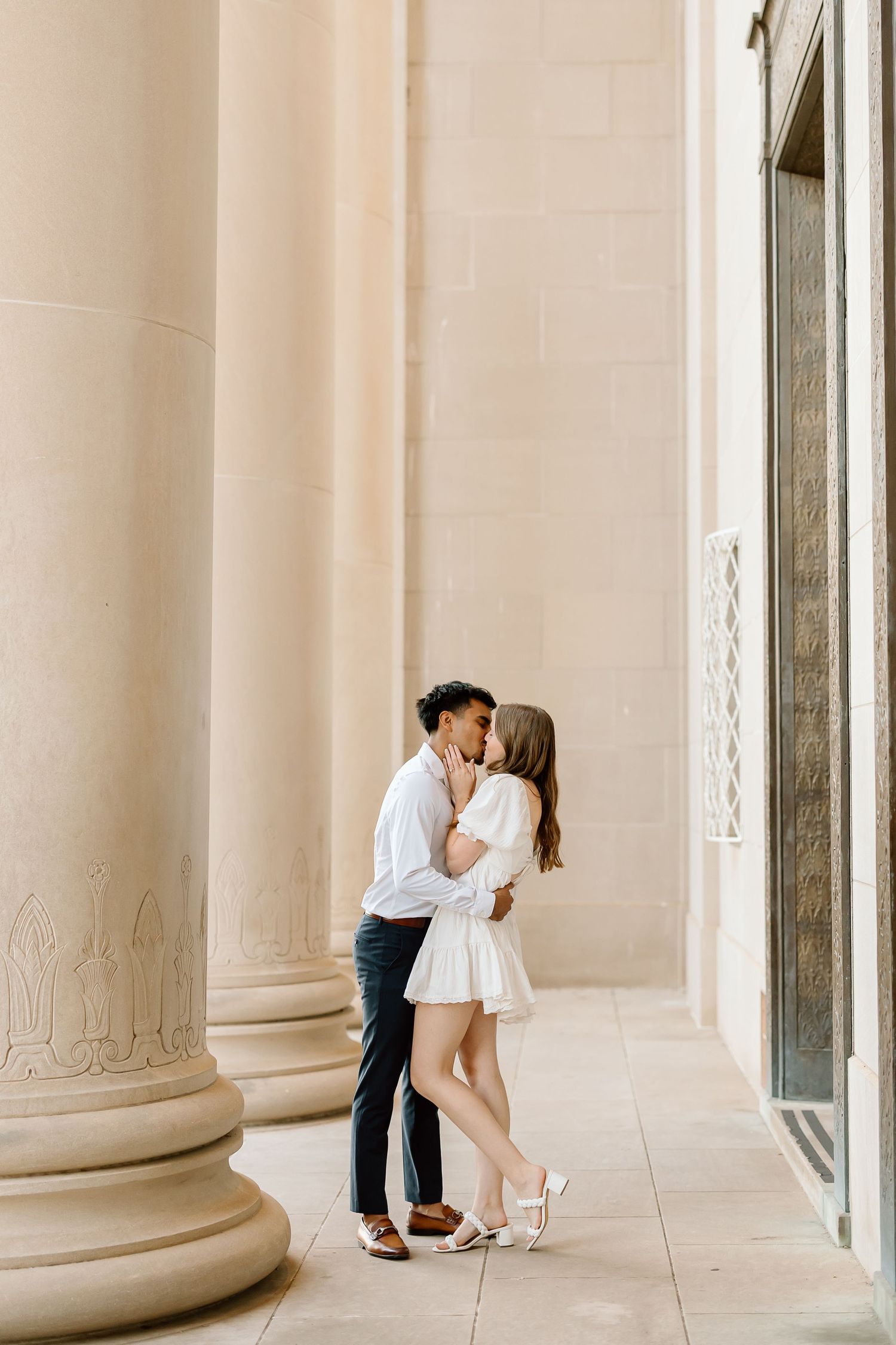 A romantic couple shares intimate moments between elegant white columns in a classical architectural setting.