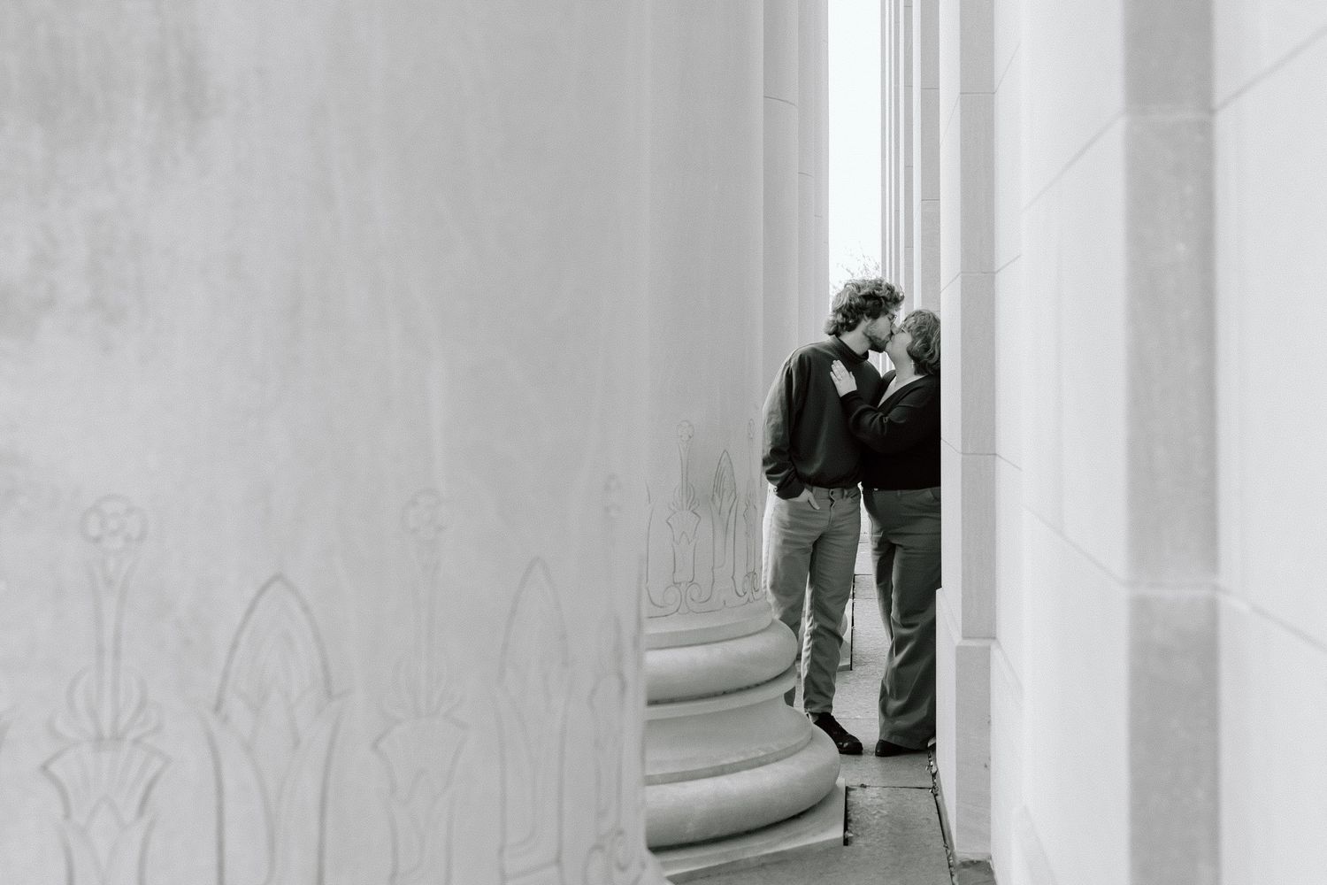 Black and white photo of couple sharing intimate moment between architectural columns.