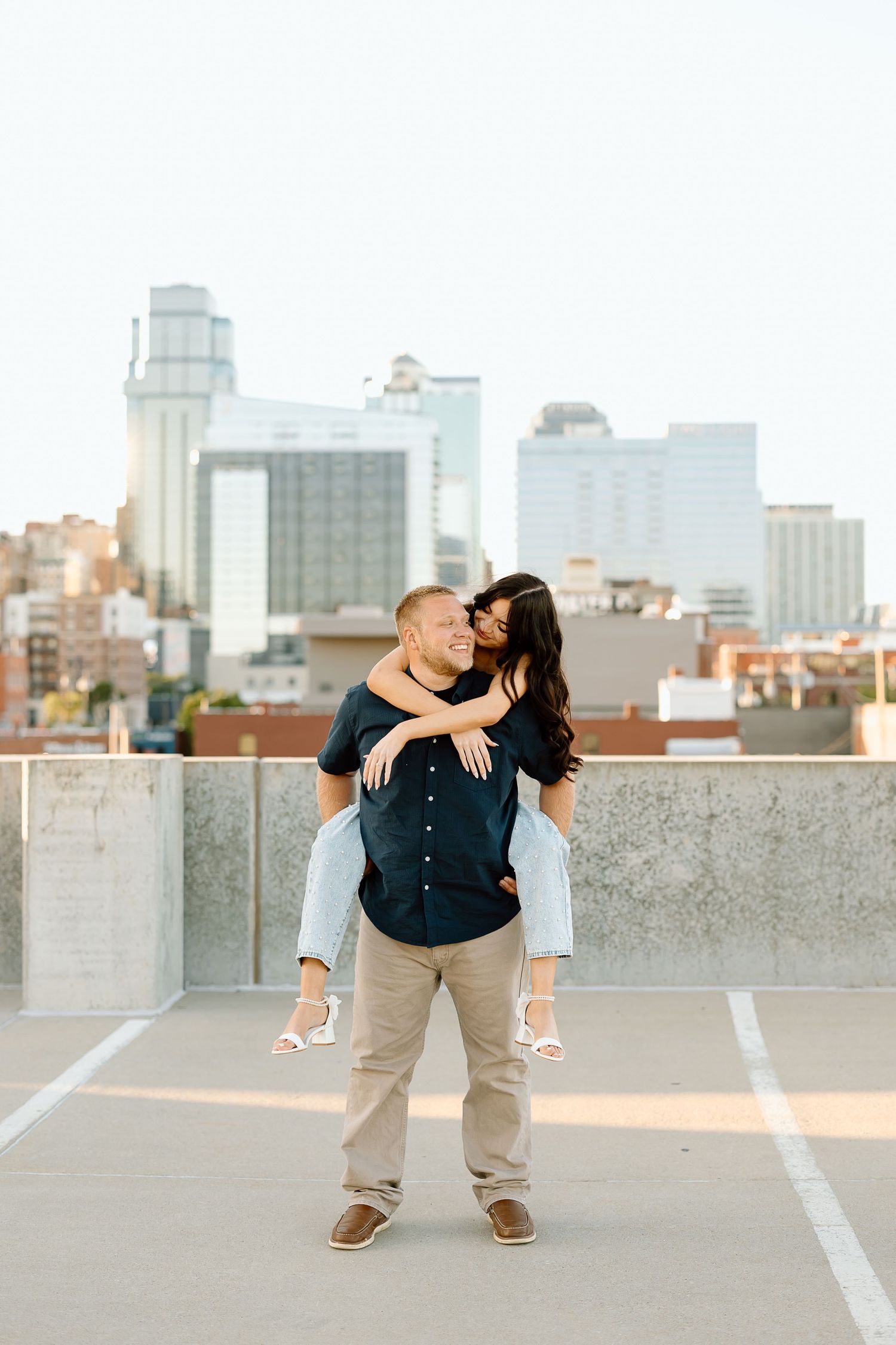 A romantic couple embraces on a rooftop parking deck with a city skyline visible in the background at sunset.