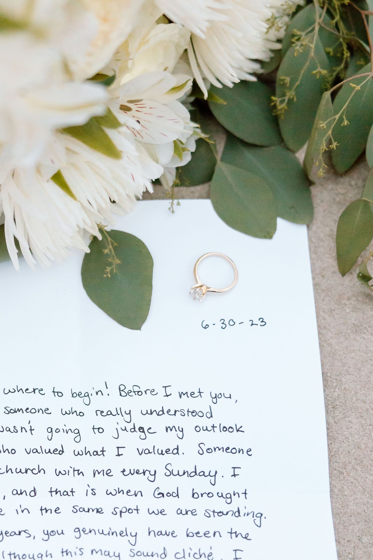 A delicate engagement ring sits on a handwritten love letter surrounded by white flowers and eucalyptus leaves.