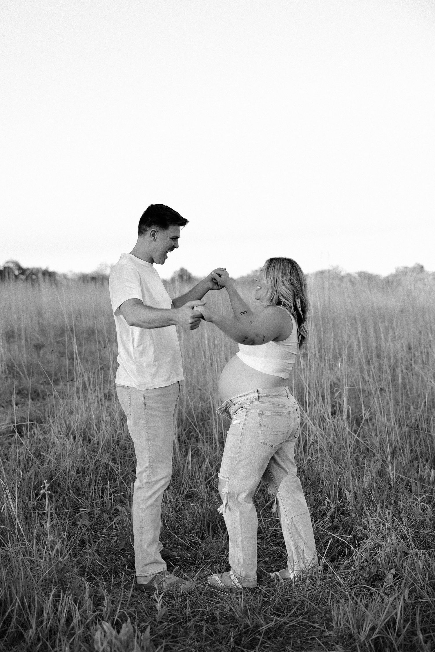 A couple in white tops and light jeans dance playfully together in a grassy field at sunset in black and white.