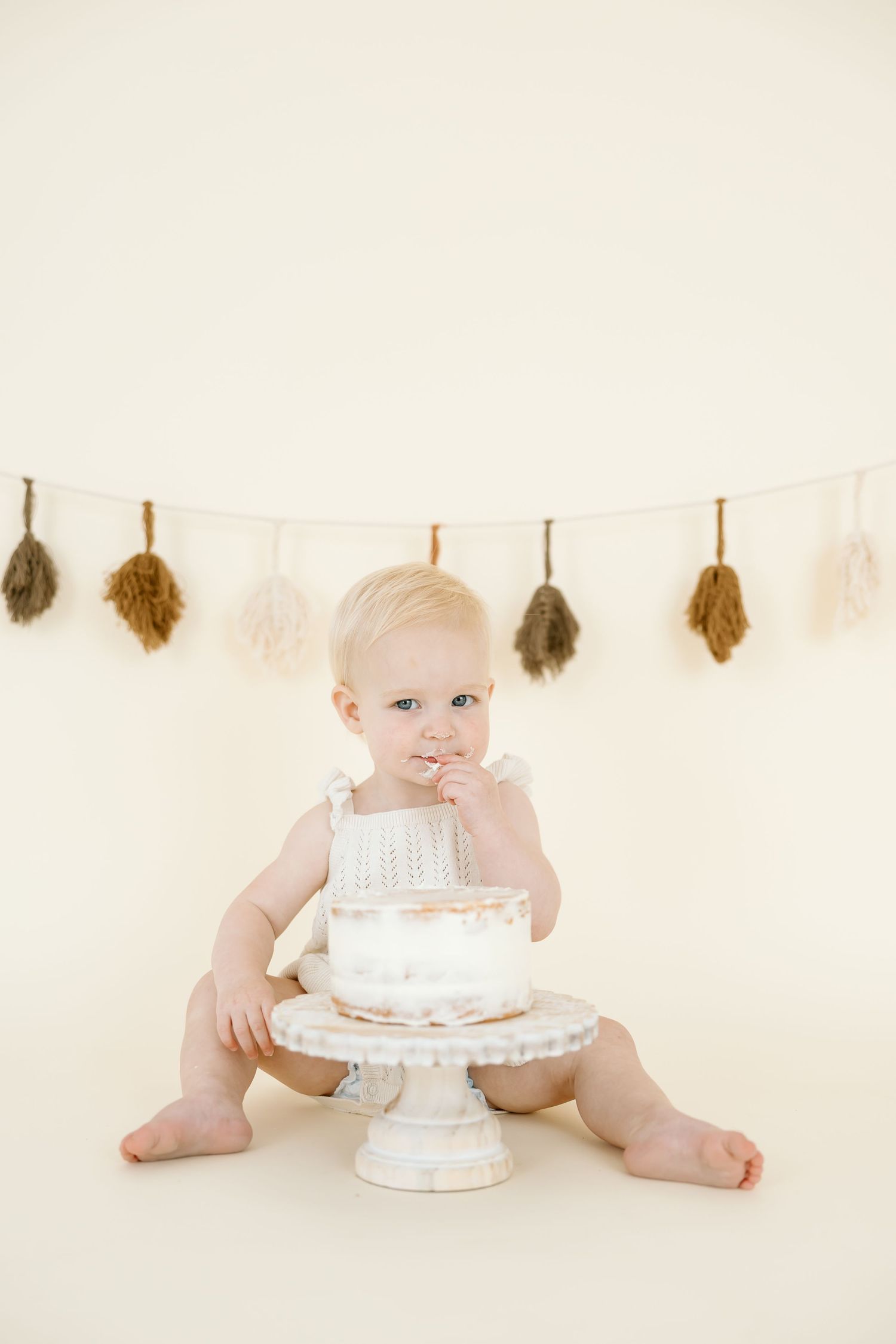 A minimalist cake smash photo series showing a baby with a white cake on a cream background with feather garland.