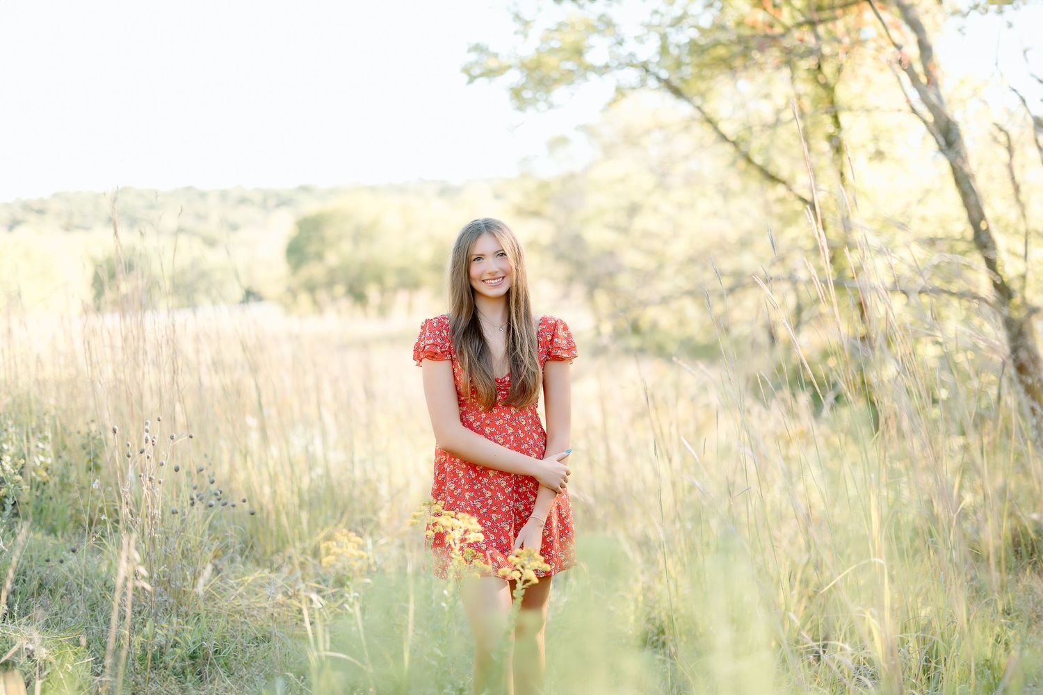 A person in a coral dress sits in a golden field at sunset with tall grass and trees in the background.