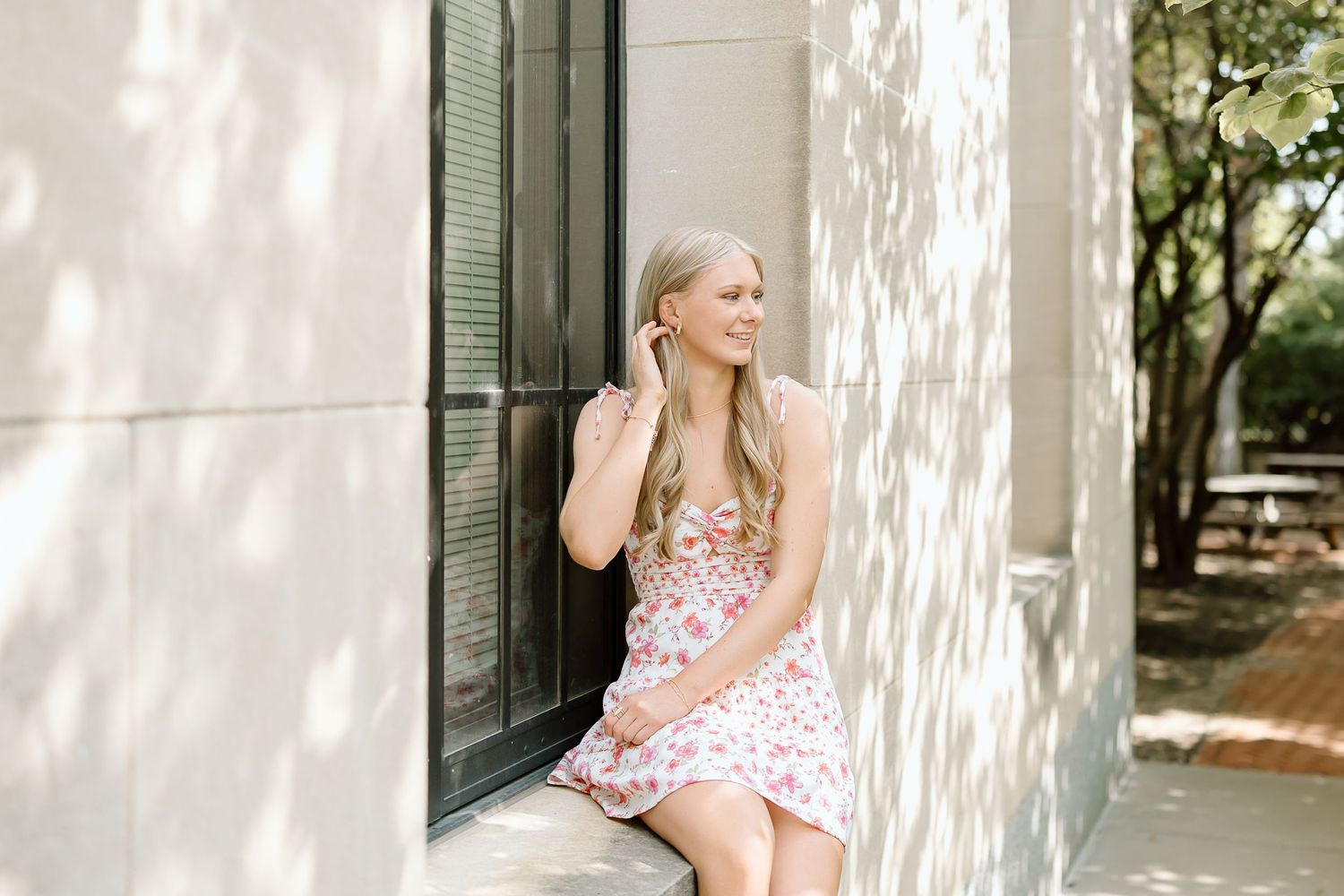 A person in a pink floral dress sits on a white porch swing in dappled sunlight near green shutters.