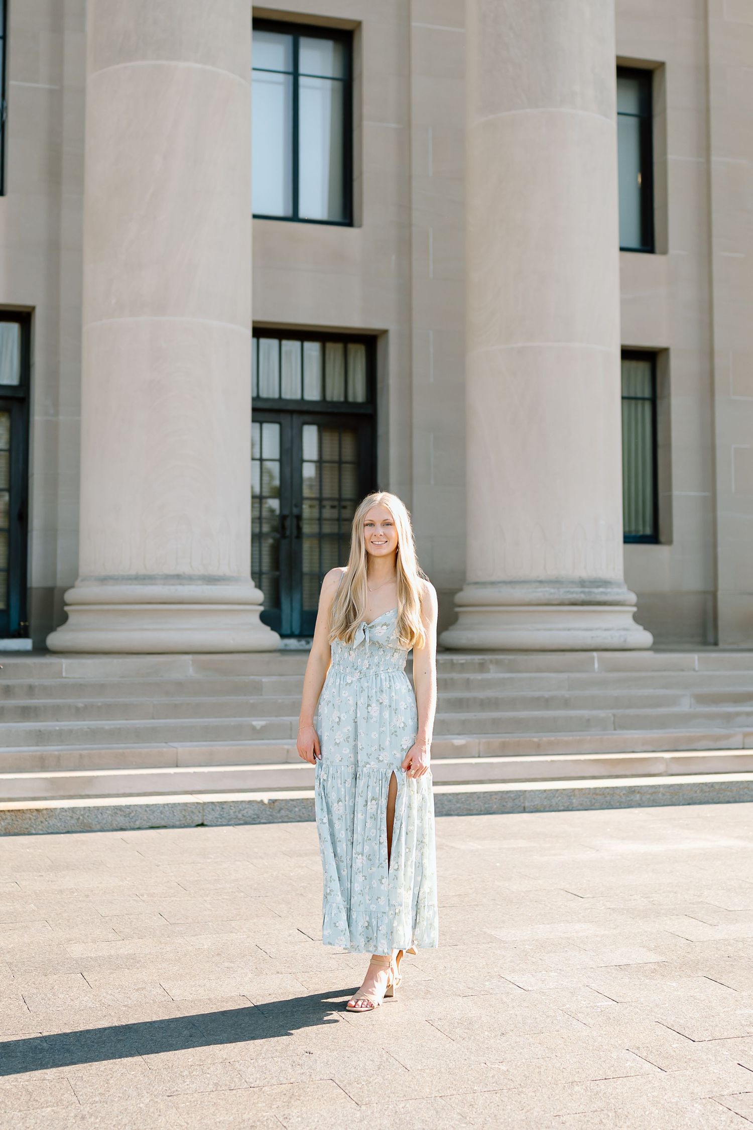 A person in a light blue floral maxi dress stands in front of a classical building with large stone columns.