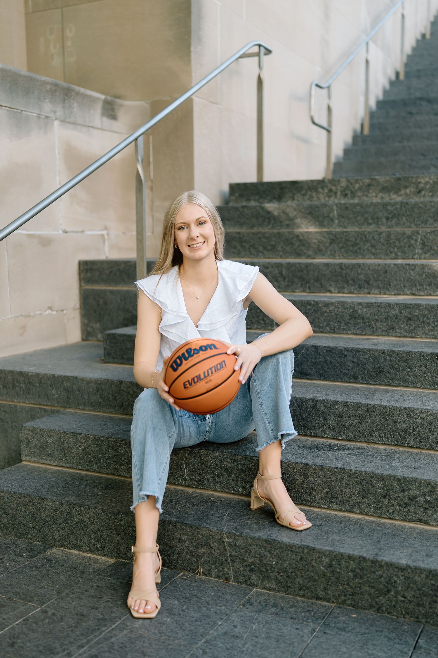 A person in a white top and ripped jeans sits on concrete steps holding a basketball and wearing sandals.