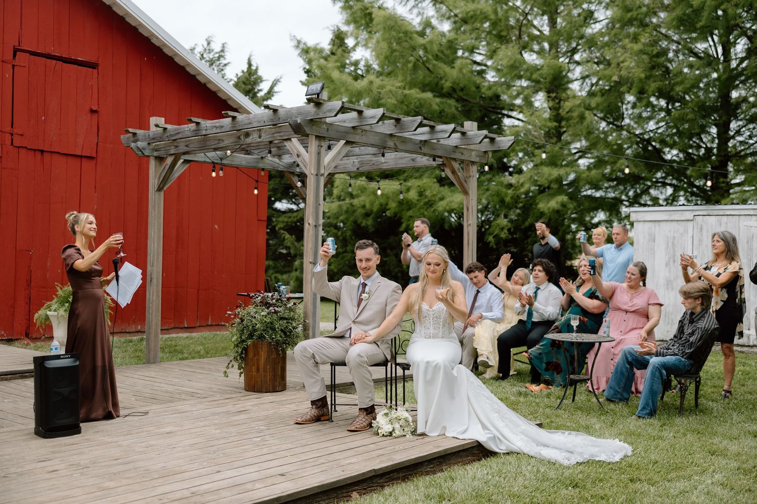 Outdoor wedding ceremony on a wooden deck with a pergola, red barn backdrop, bride and groom seated while guests applaud.