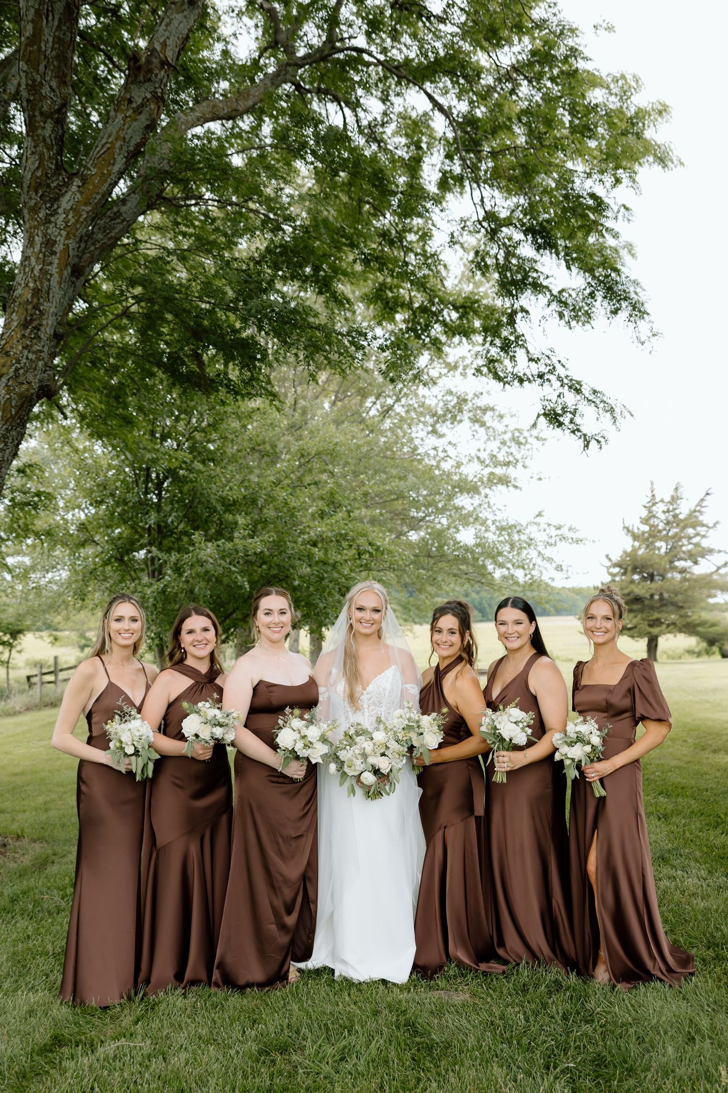 Bride in white gown with six bridesmaids in brown satin dresses holding white bouquets outdoors under a large tree.