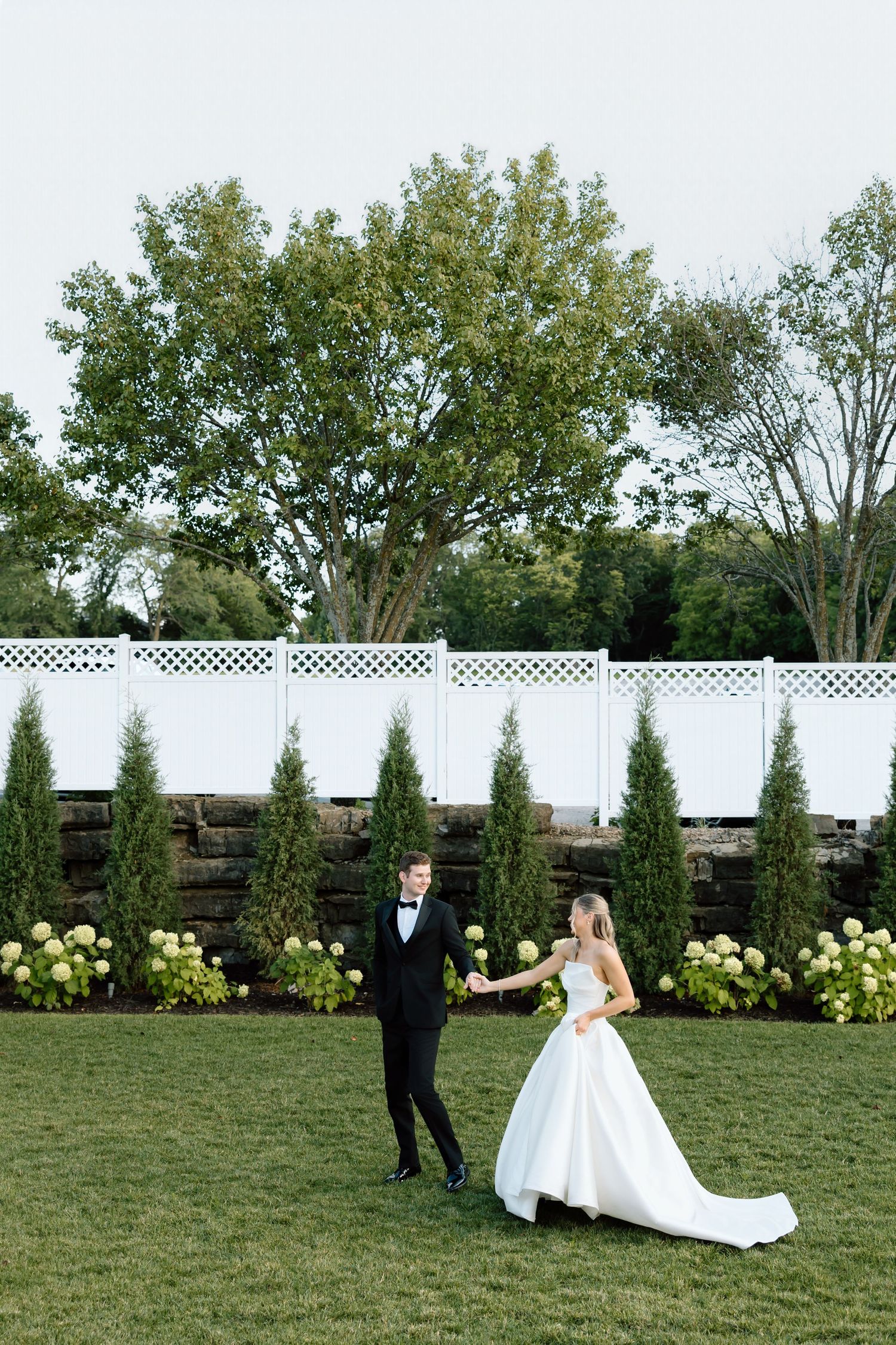 A bride and groom dance on a lush green lawn surrounded by tall evergreens and hydrangeas at an elegant outdoor wedding.