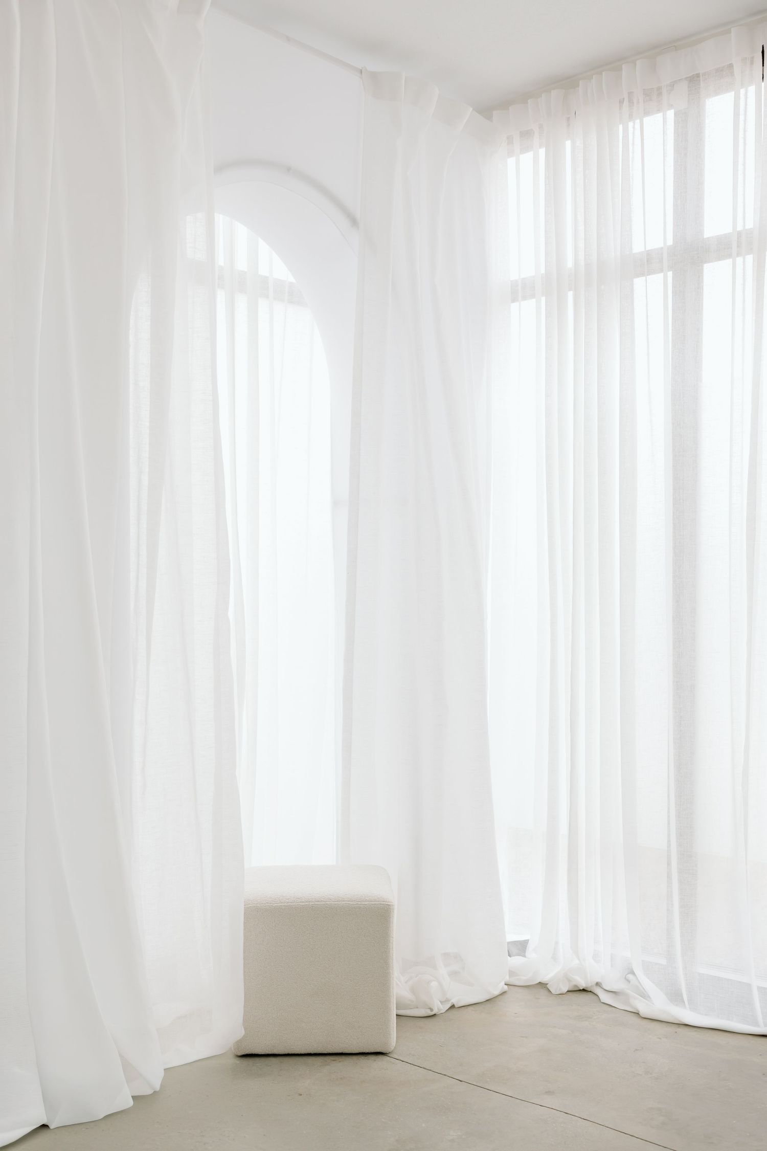 Minimalist white room interior with flowing sheer curtains and a single cream-colored ottoman against bright windows.