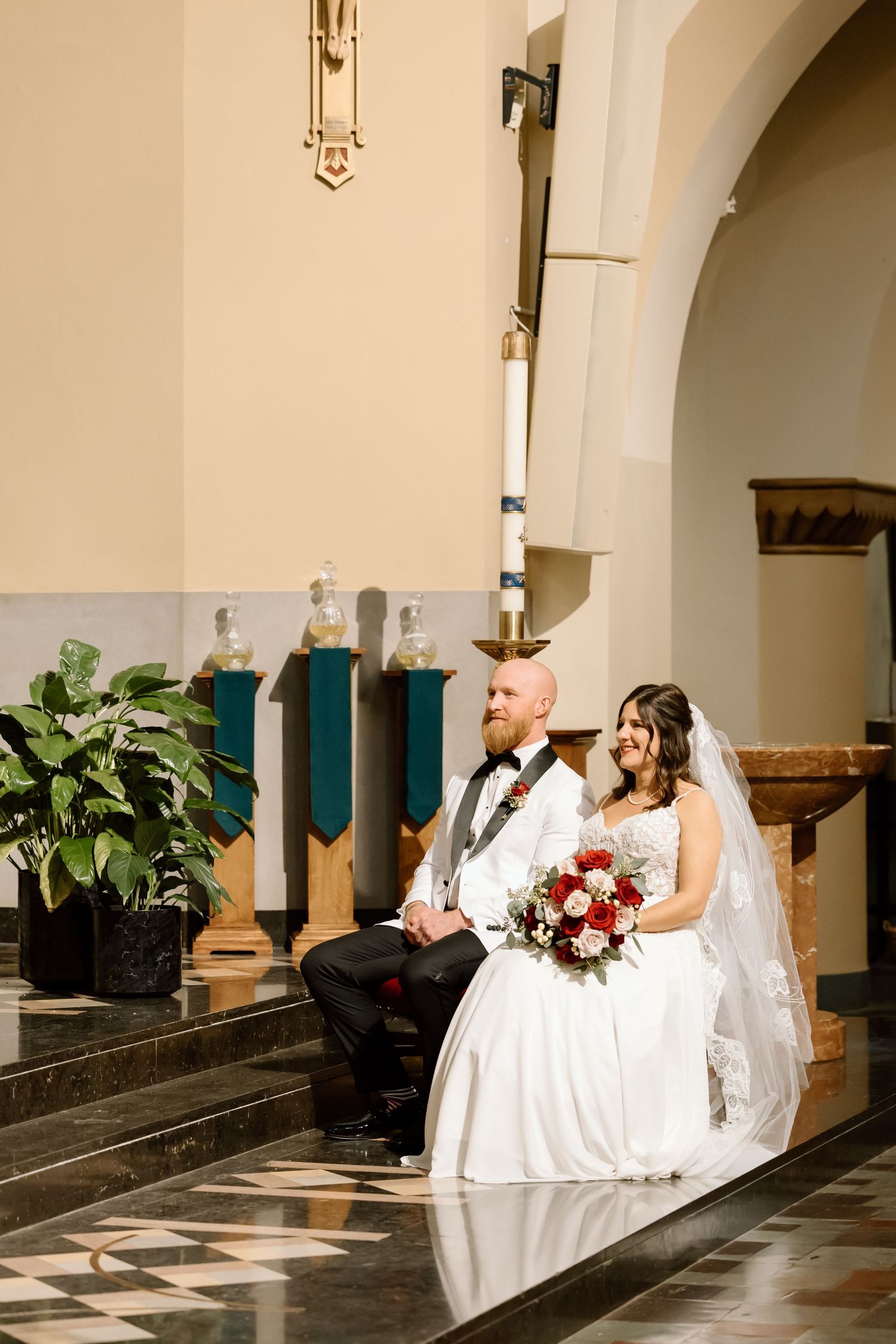 A bride and groom sit together on church altar steps with green plants and decorative arches in the background.