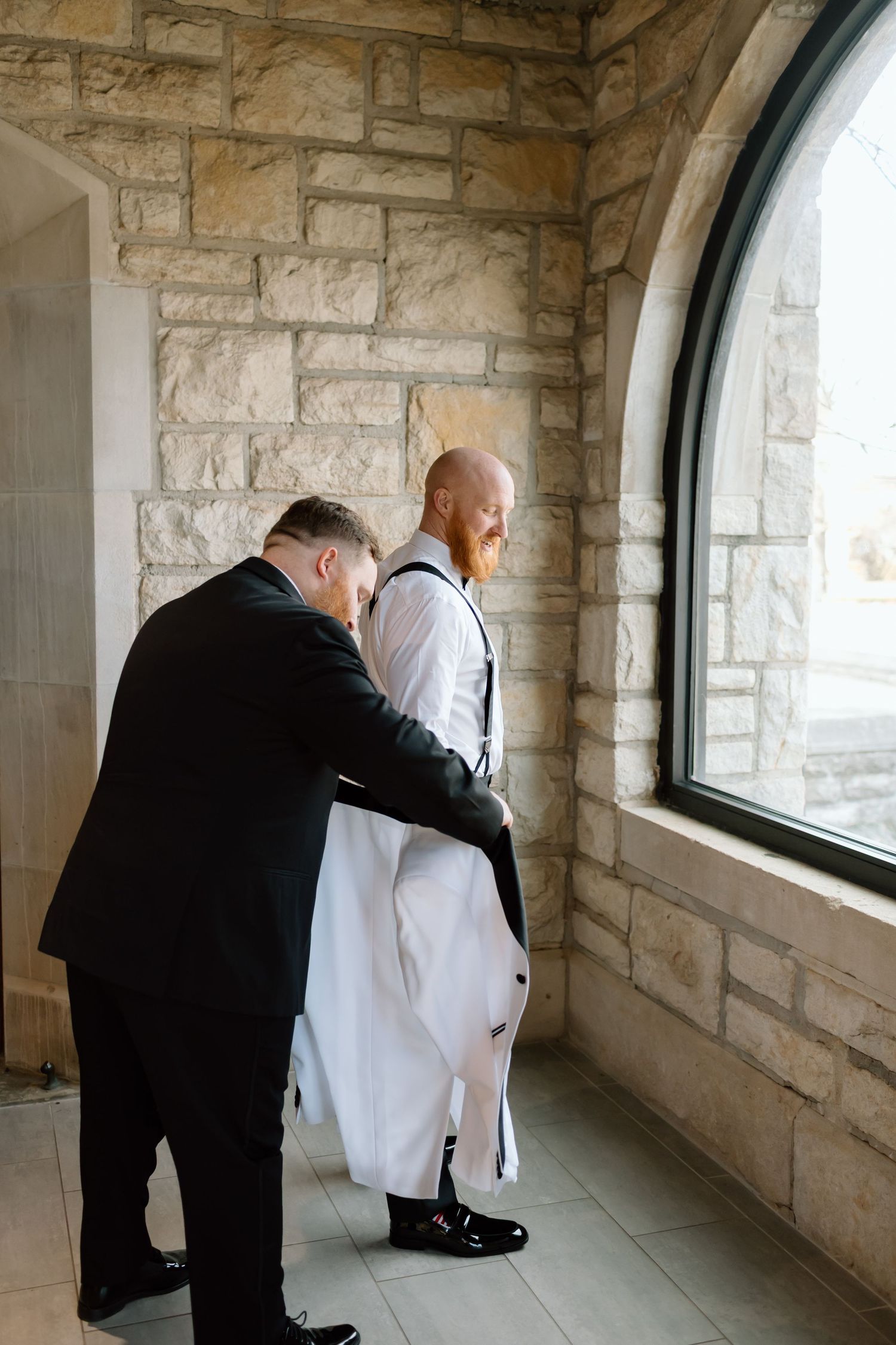 Two people in formal attire lean towards a window inside a stone-walled corridor.