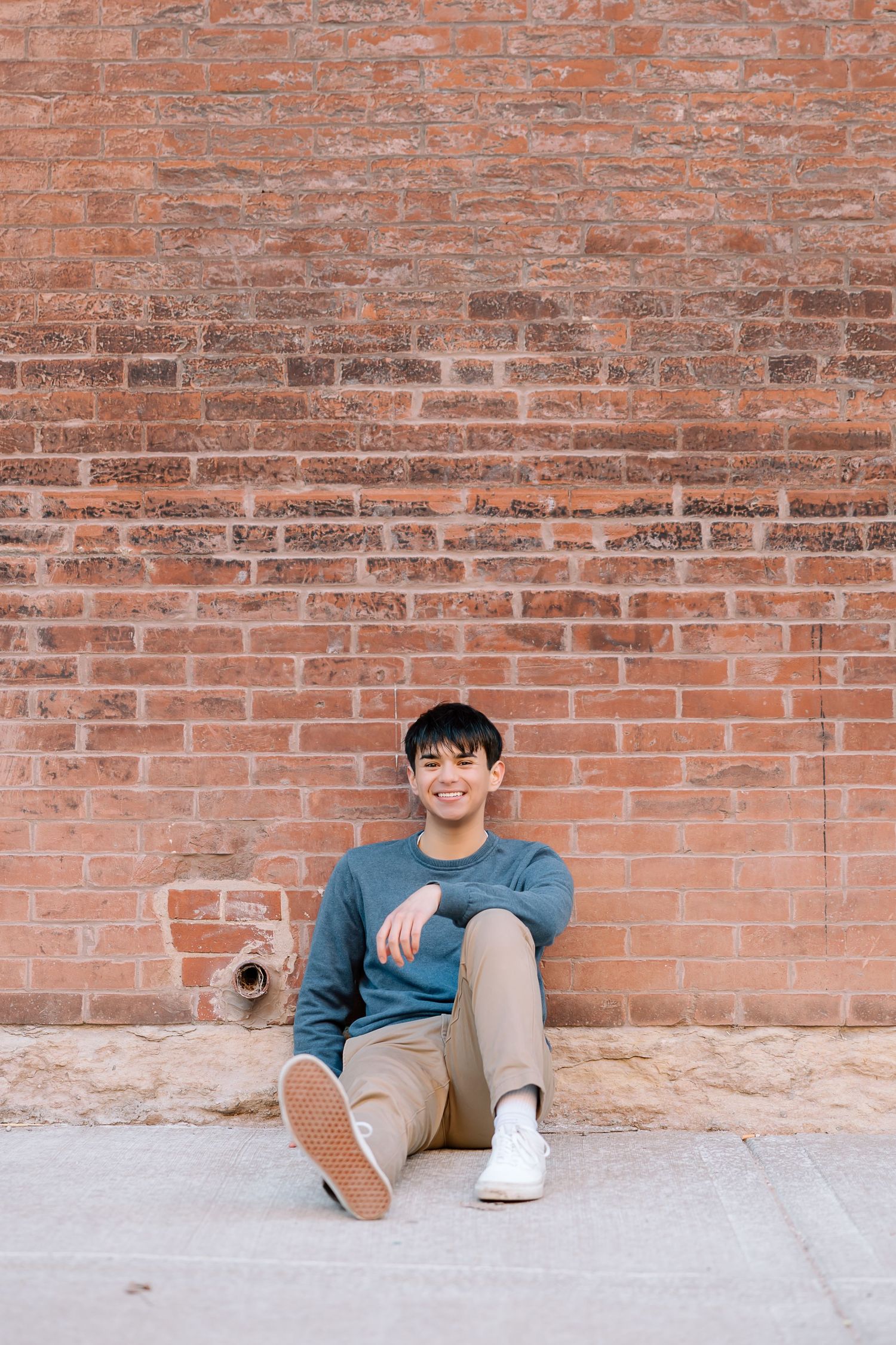 Person in blue sweater and khaki pants sitting casually against rustic brick wall.