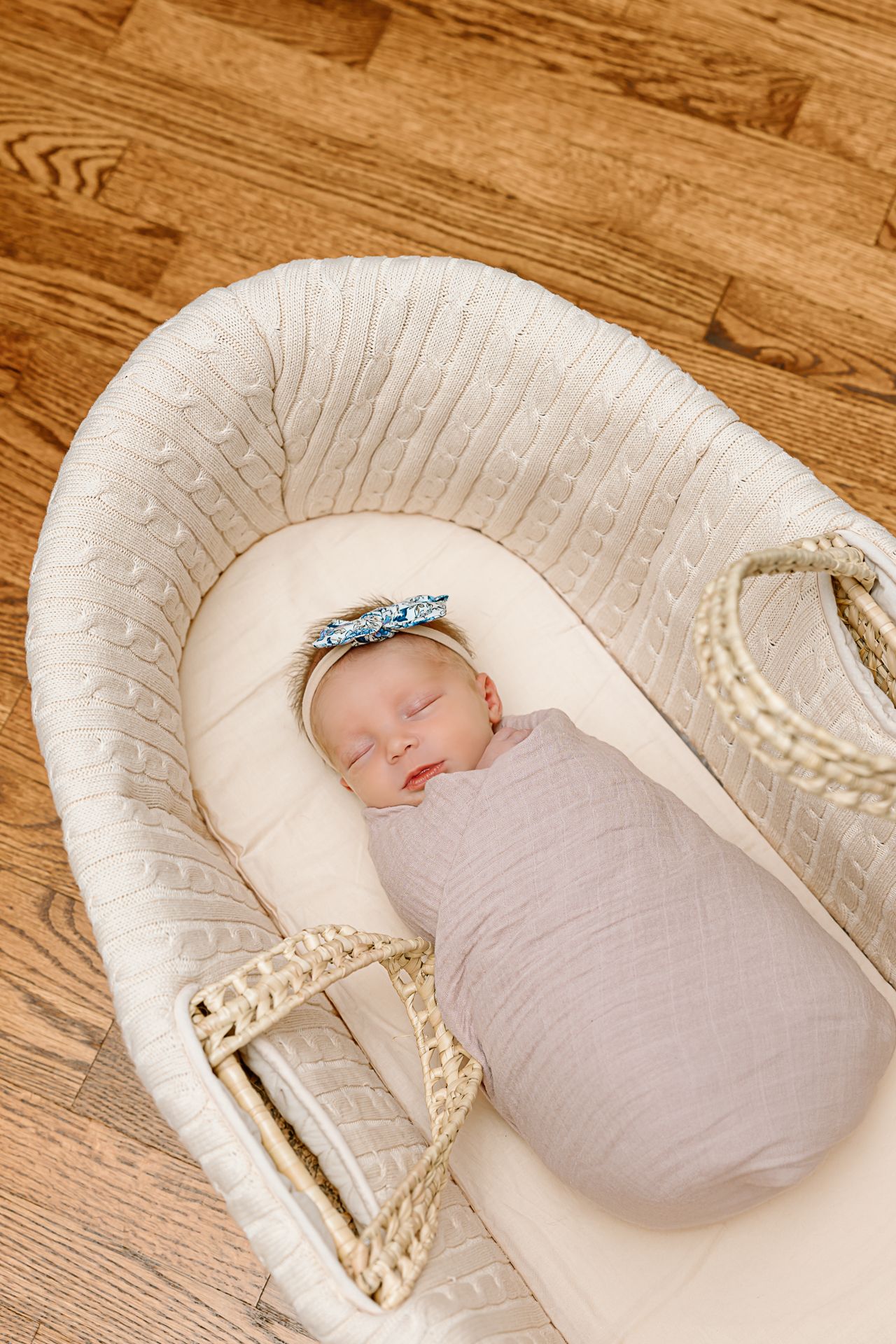 Overhead view of sleeping newborn in white bassinet on hardwood floor with decorative details.