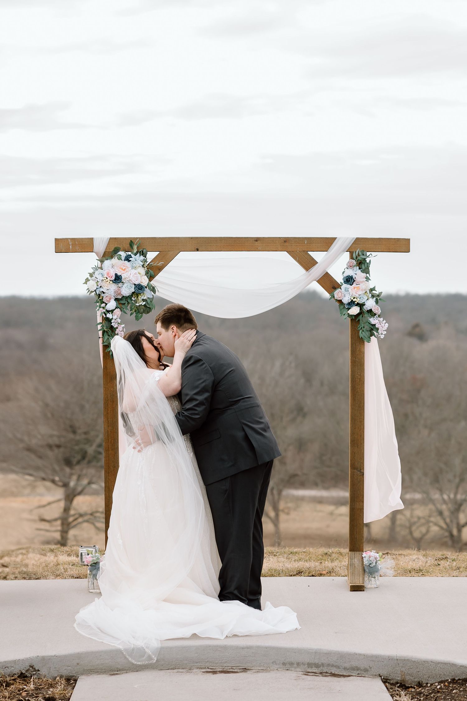 Newlyweds sharing first kiss under wooden arch decorated with white fabric and flowers.