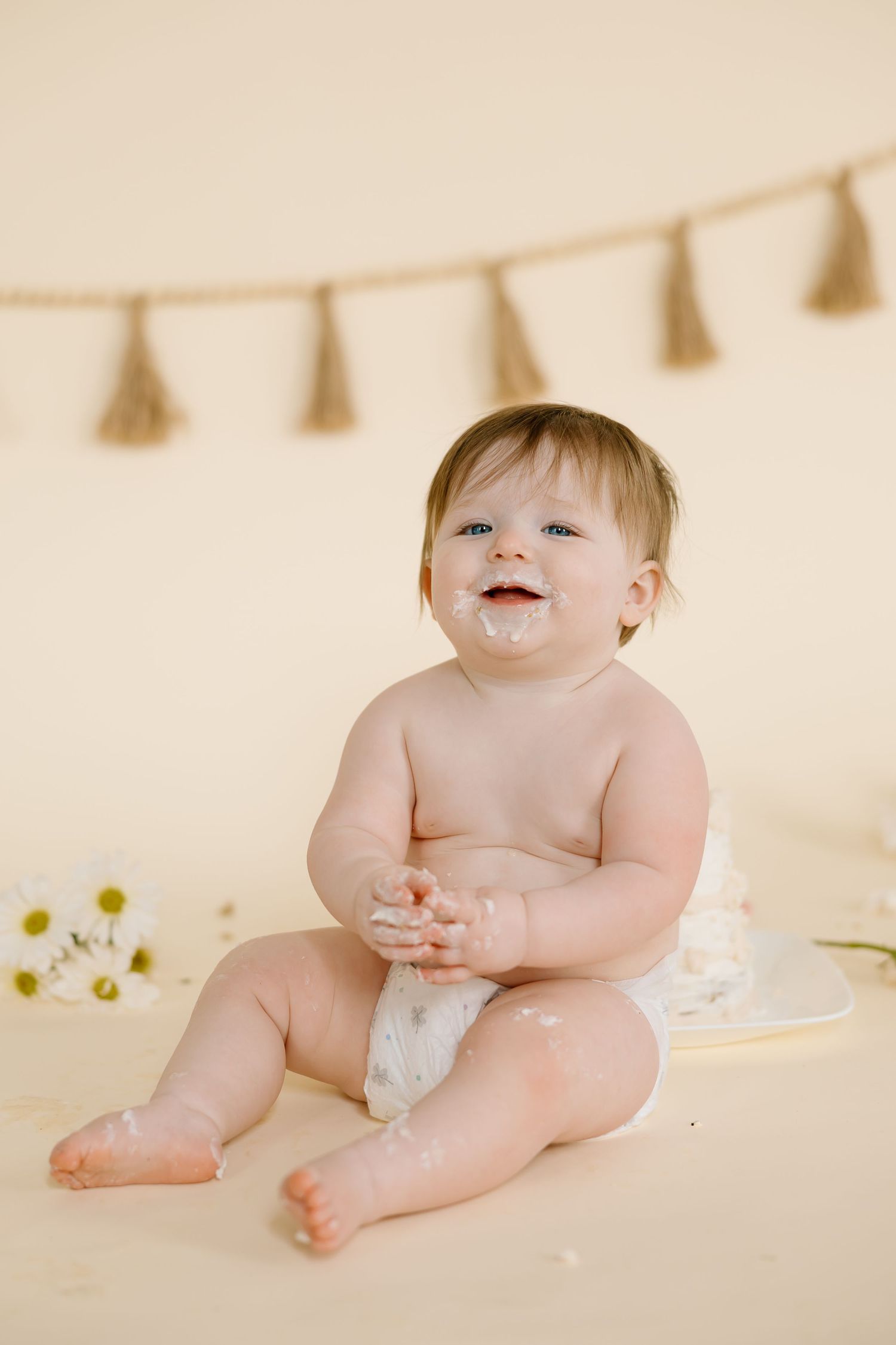 Happy baby sits with decorative tassels and flowers during milestone photo session.