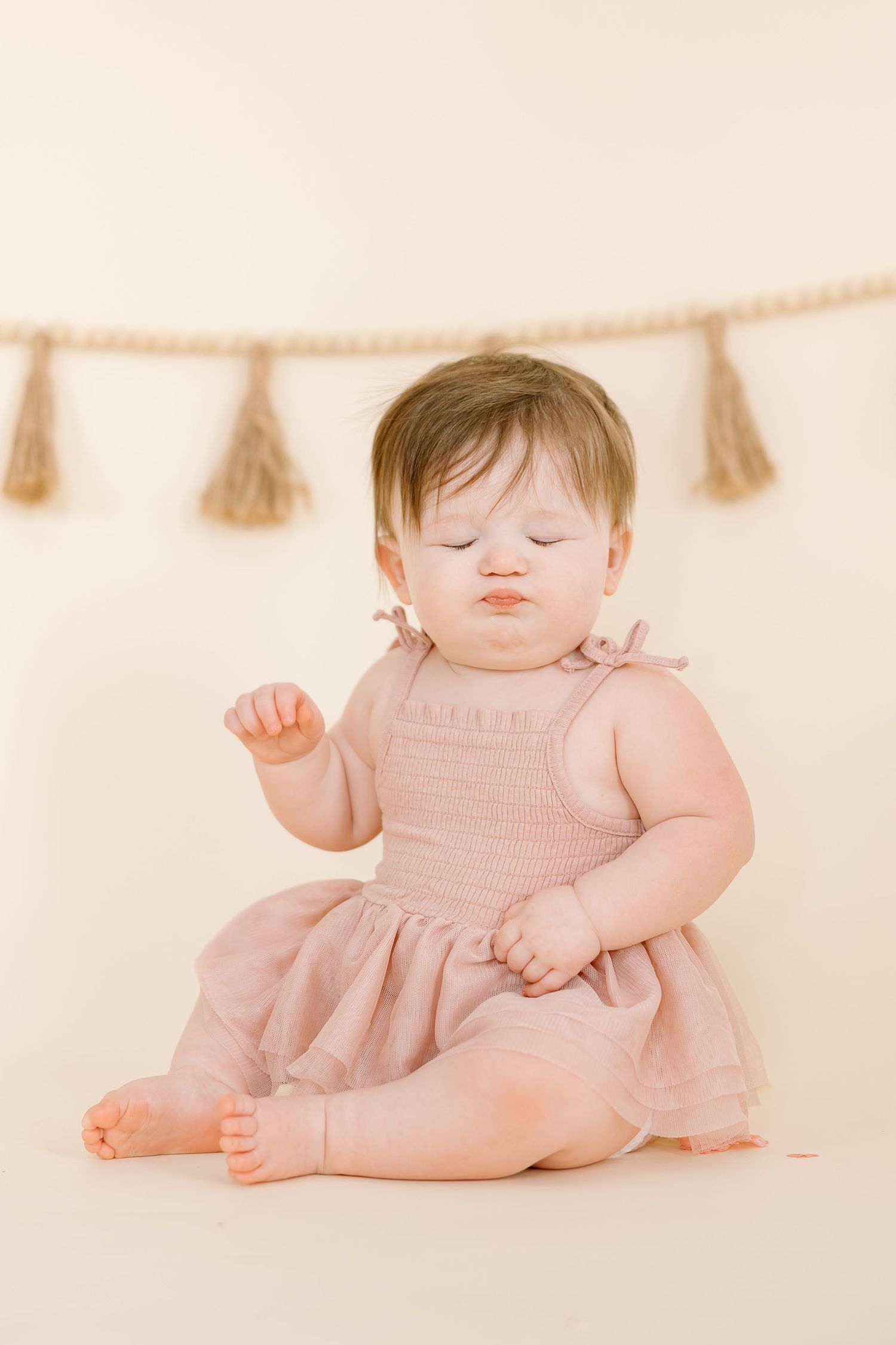 Baby in pink dress sits on floor with eyes closed under hanging tassel garland.