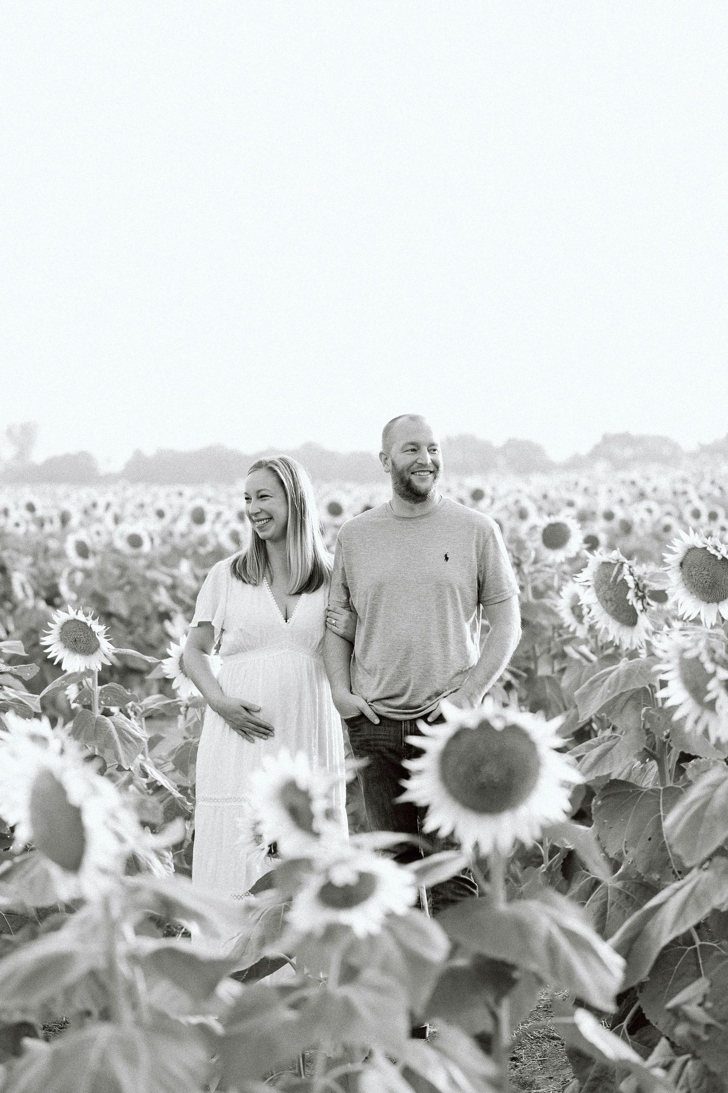 A couple stands together in a vast field of blooming sunflowers on a bright summer day.