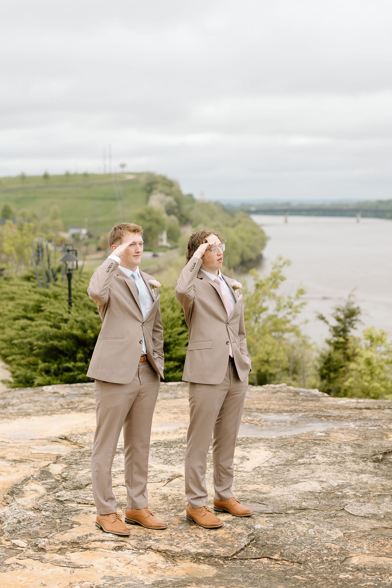 Two people in matching tan suits and brown shoes stand together on a rocky overlook with a scenic lake view.