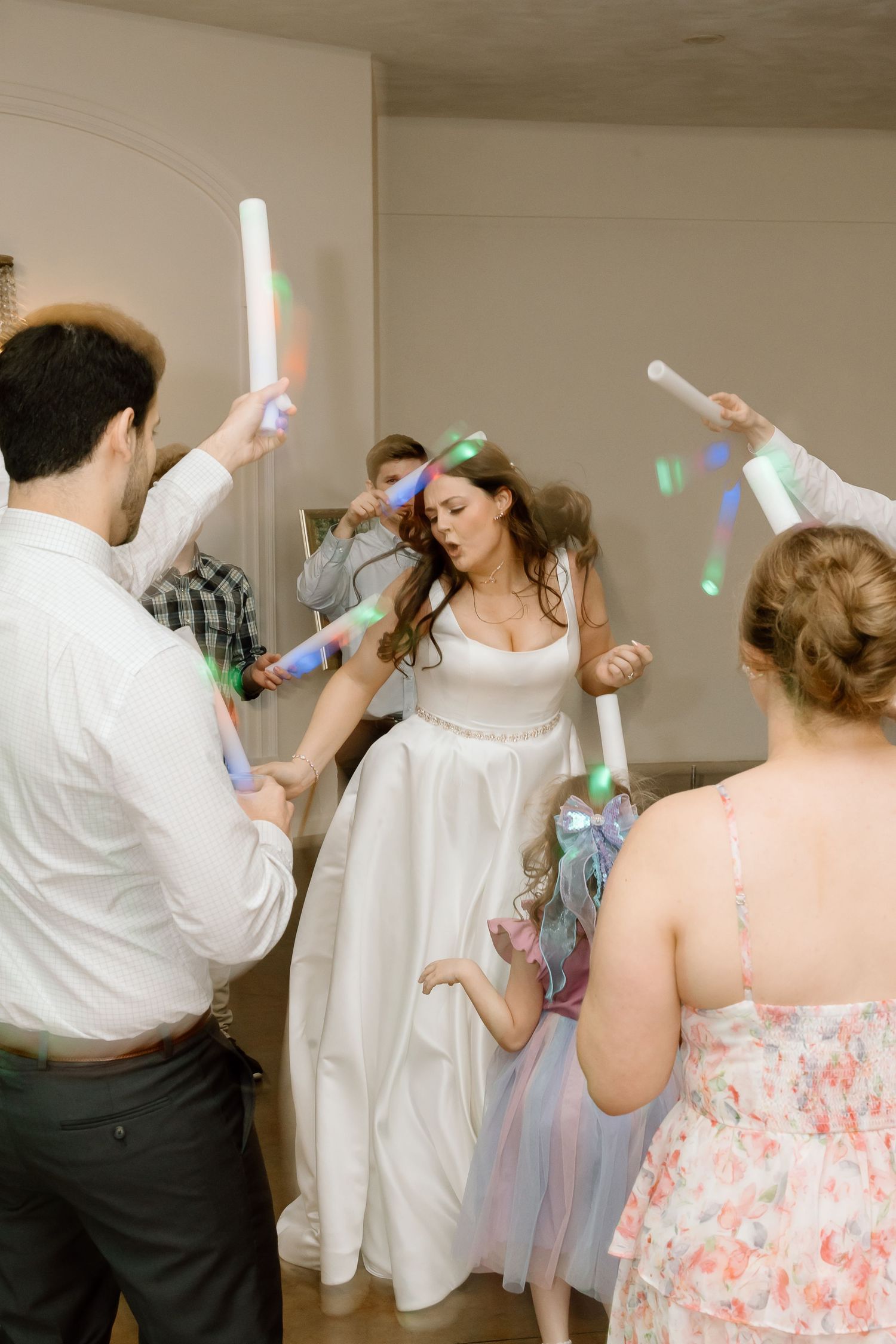 Wedding celebration with people dancing and waving light-up foam sticks at an indoor reception party.