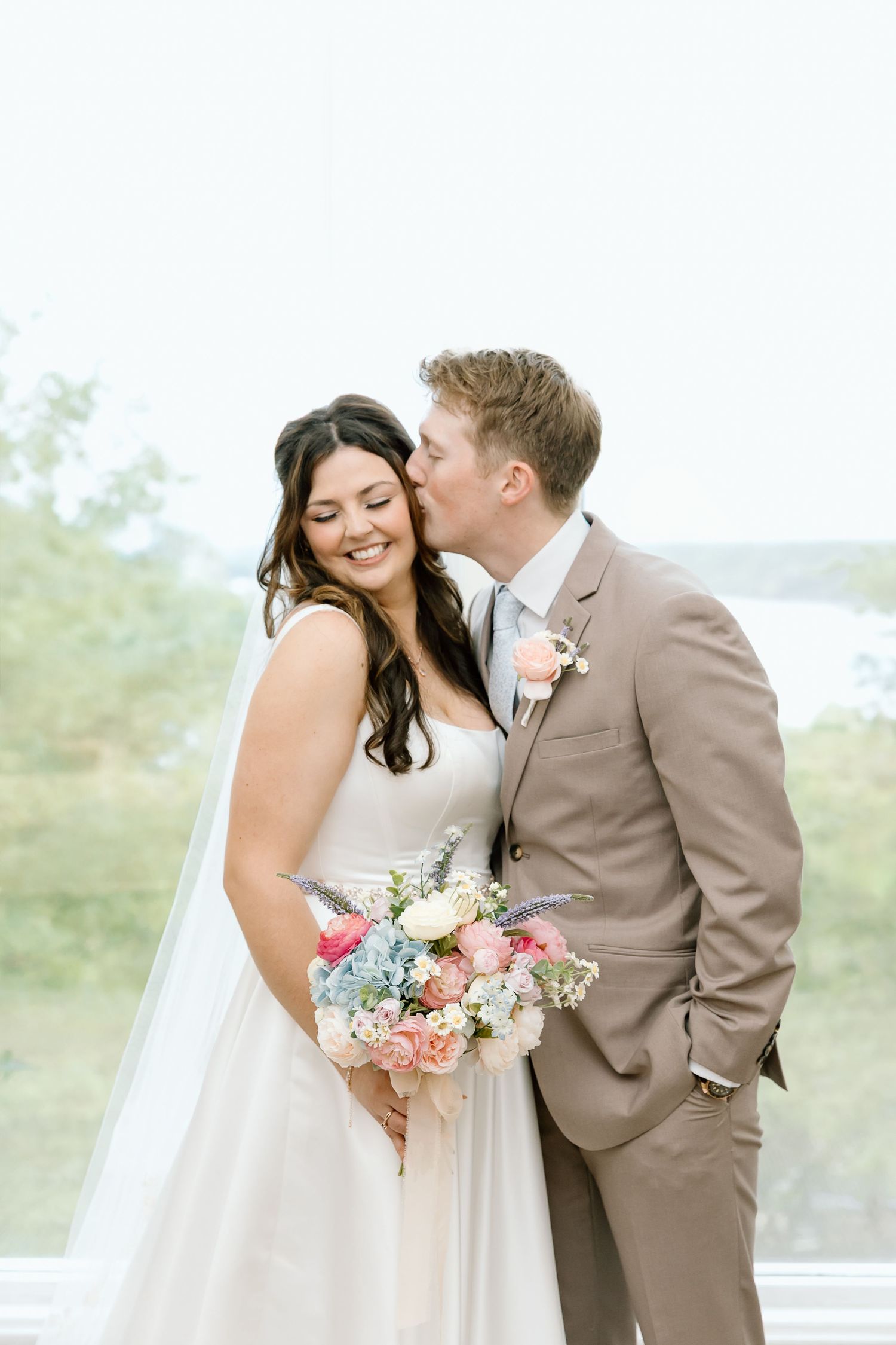 A romantic wedding couple shares tender moments in a light tan suit and white dress while holding a pink floral bouquet.