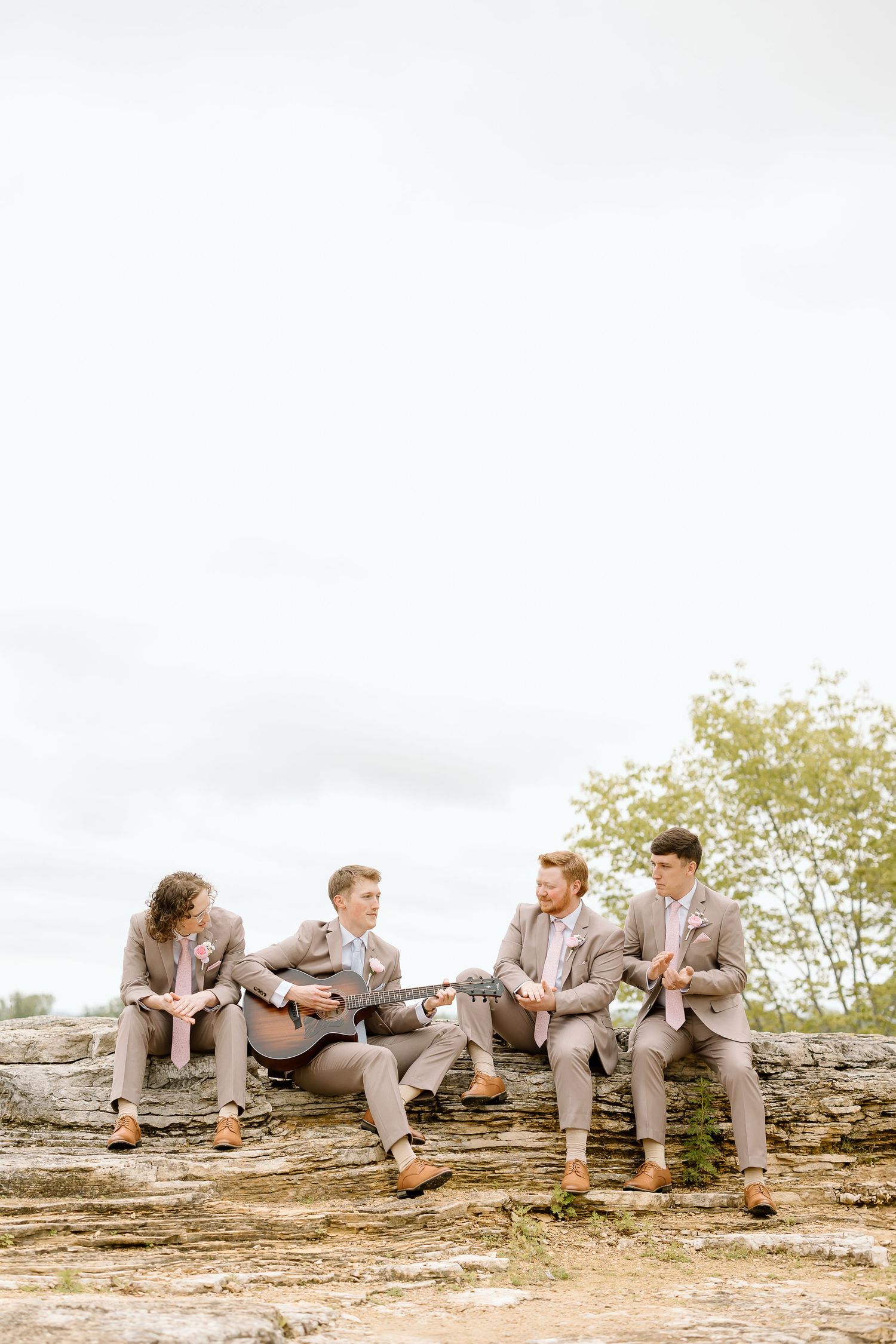 Groomsmen in gray suits sit casually on a rustic stone wall outdoors during a wedding photoshoot.