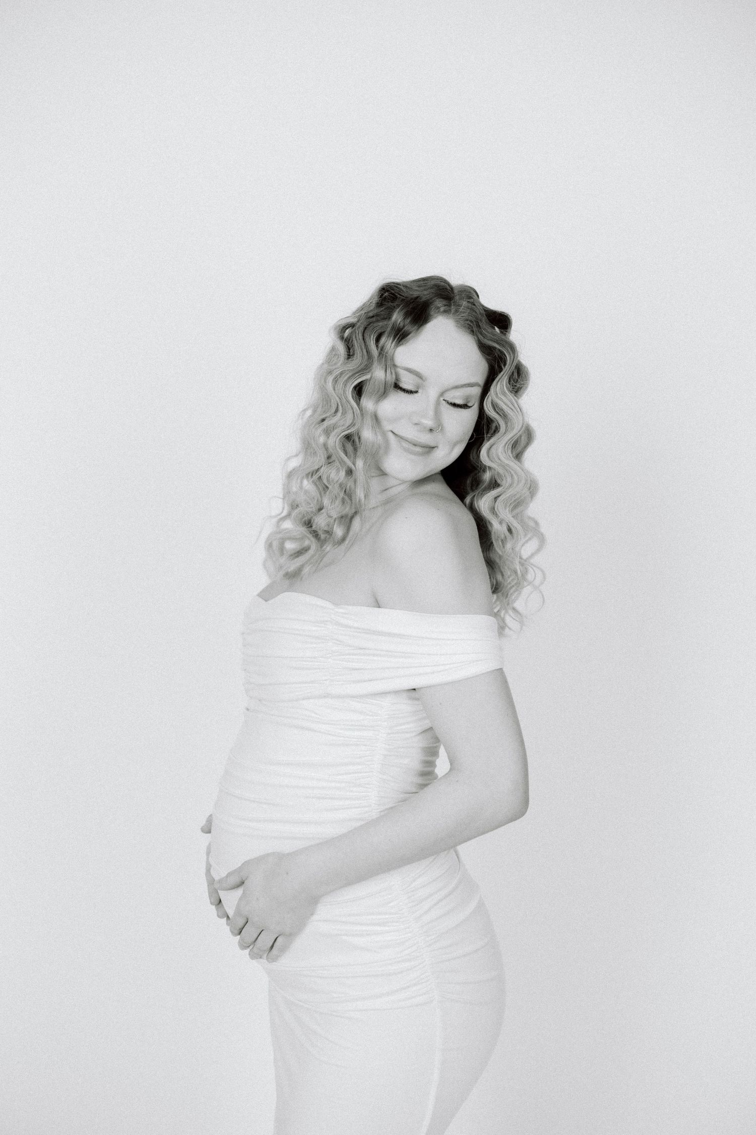 Black and white maternity photo series showing woman in white dress with curly hair against plain backdrop.