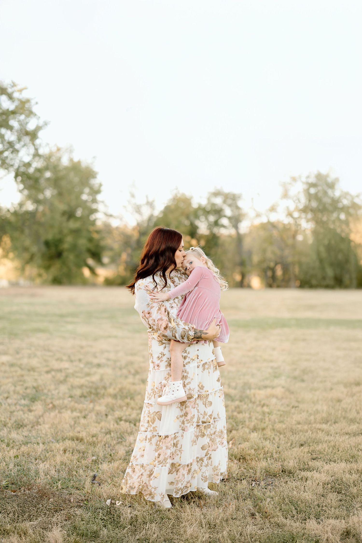 A couple in floral dress and pink shirt embrace in a golden sunlit field during sunset.
