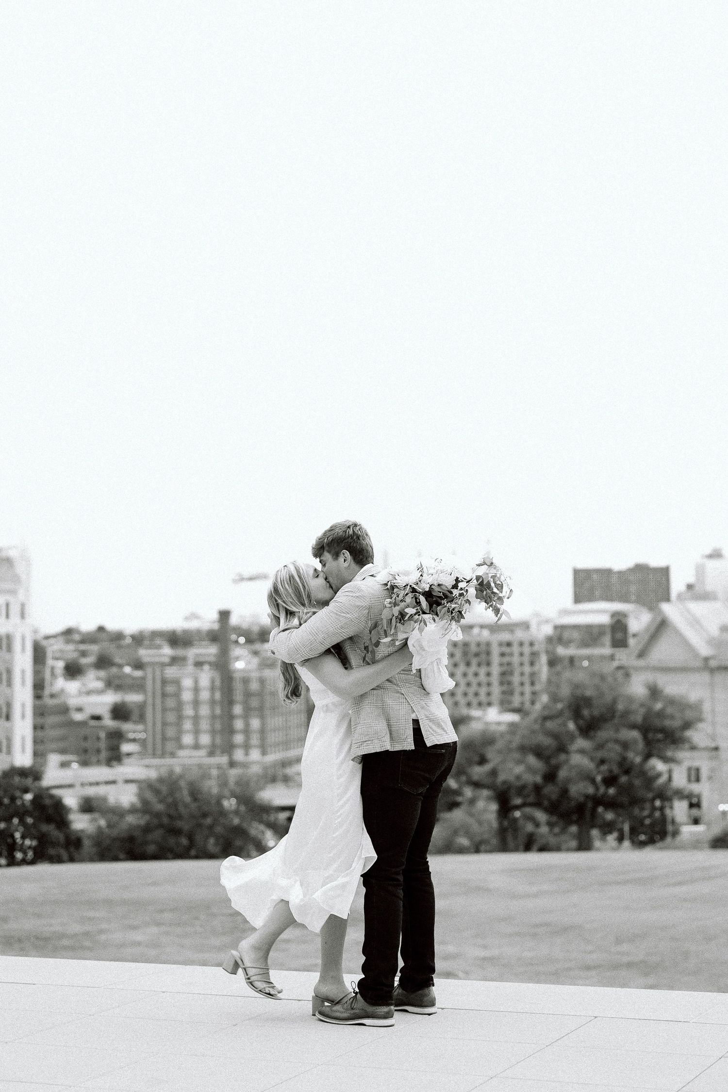 A couple in wedding attire embraces and dances together on an outdoor terrace with a city skyline in the background.