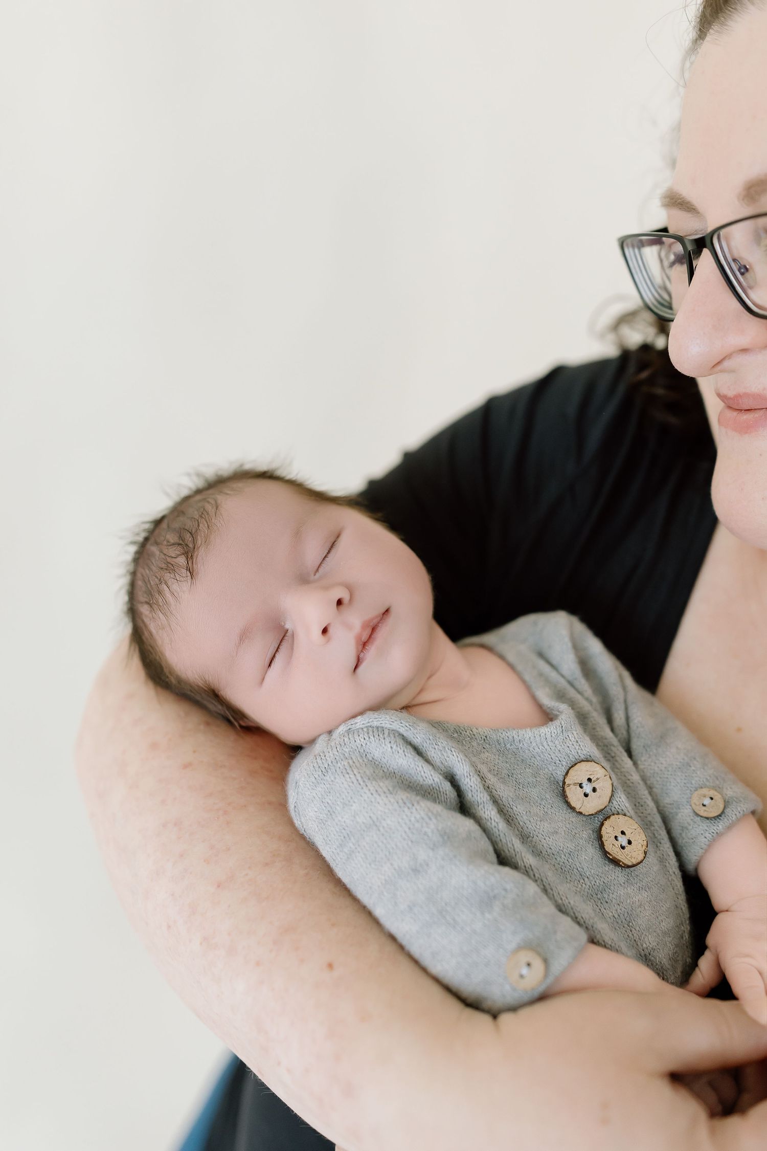 A sleeping baby wearing a gray onesie with wooden buttons rests peacefully against a caregiver's shoulder.