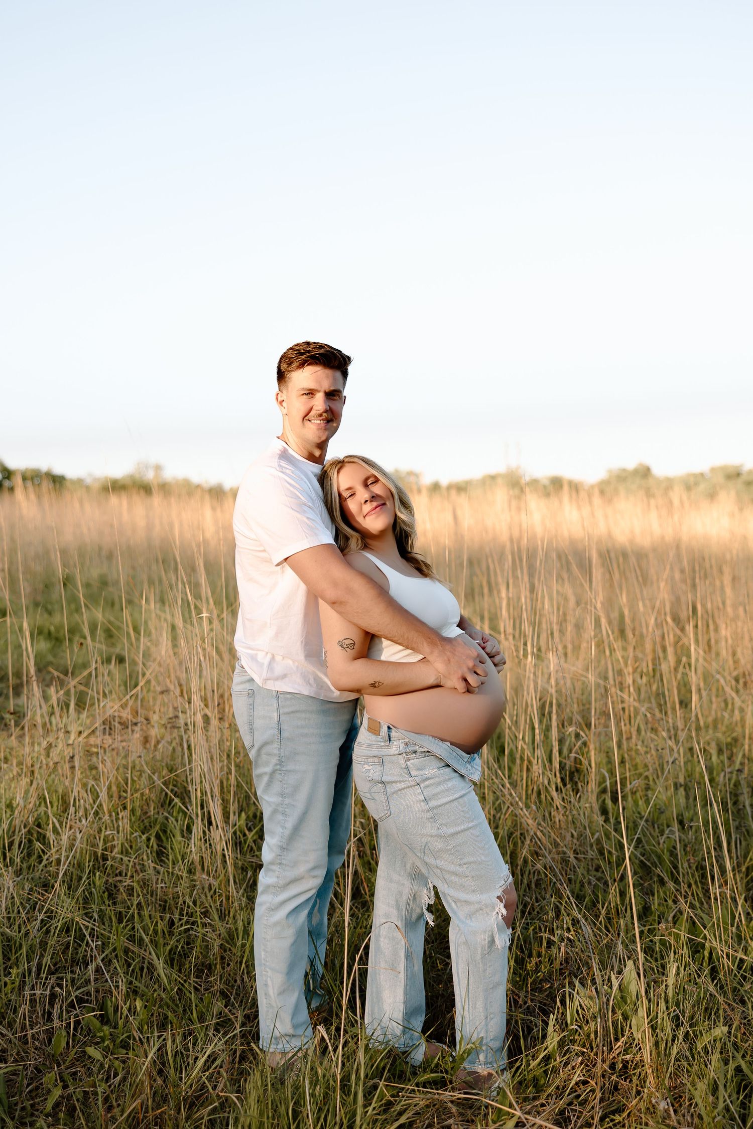 A couple embraces in a golden field at sunset as they pose for a maternity photo.