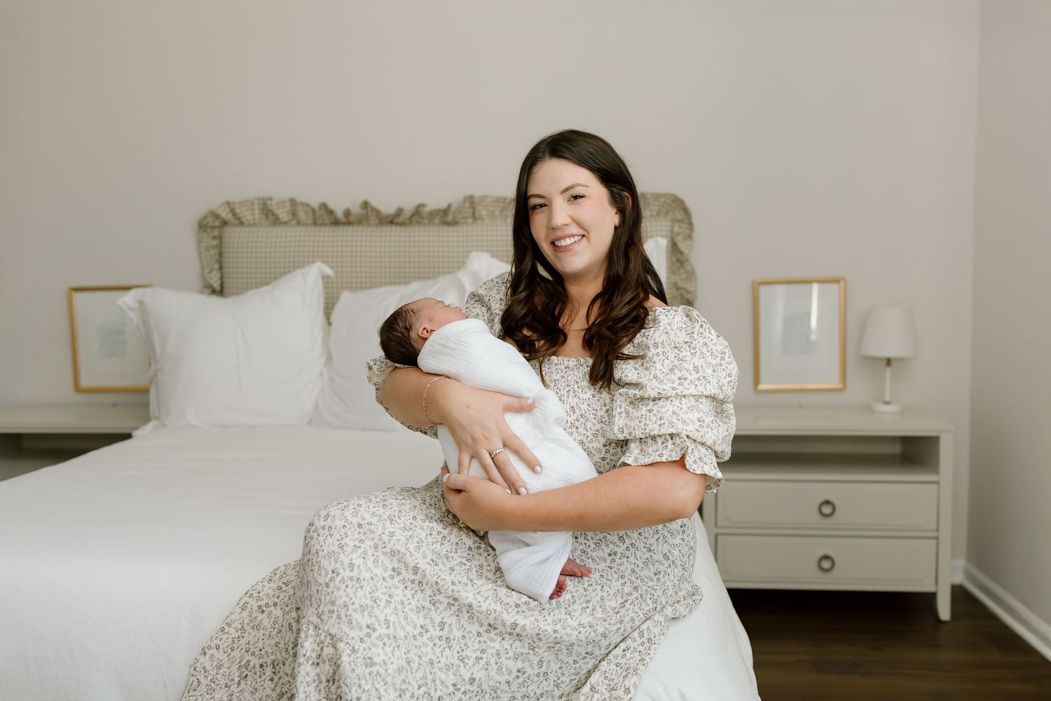 A person in a floral dress sits on a white bed holding a newborn baby wrapped in swaddling cloth.