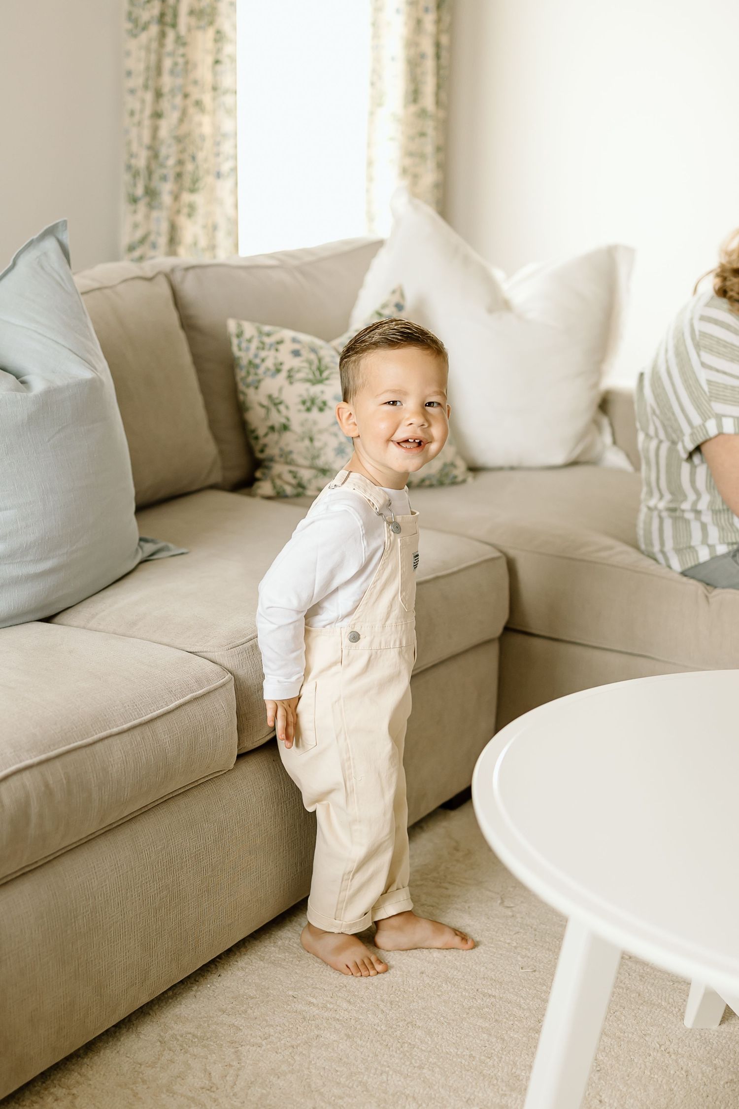 A toddler in white clothing stands and smiles while playing near a beige couch in a bright, airy living room.