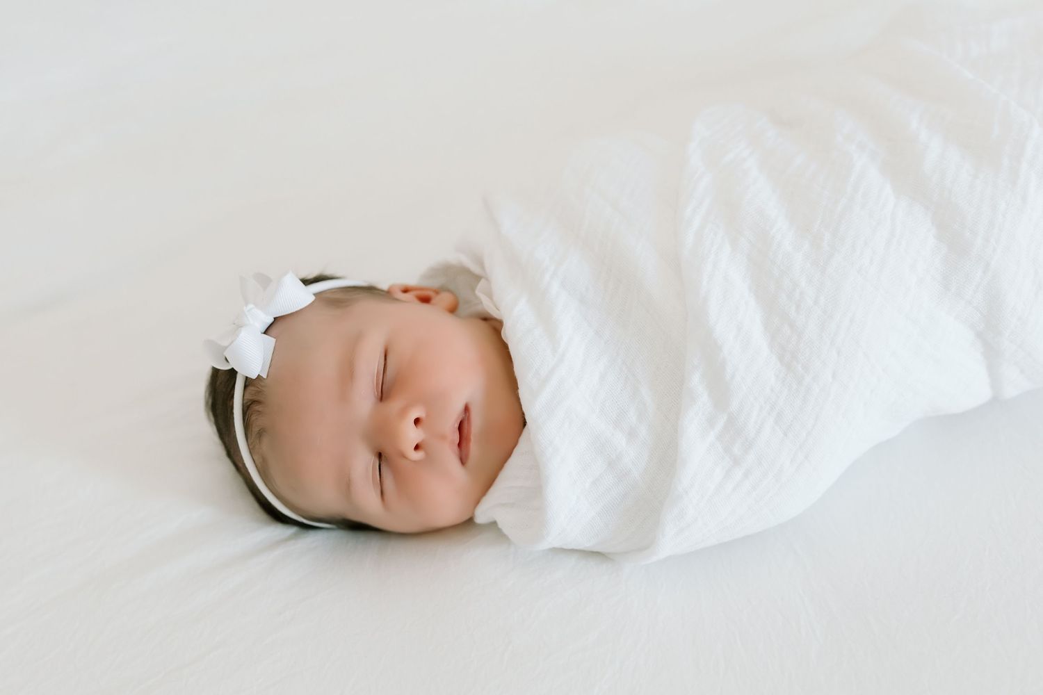 A sleeping baby wrapped in soft white swaddling blankets lies peacefully on white bedding.