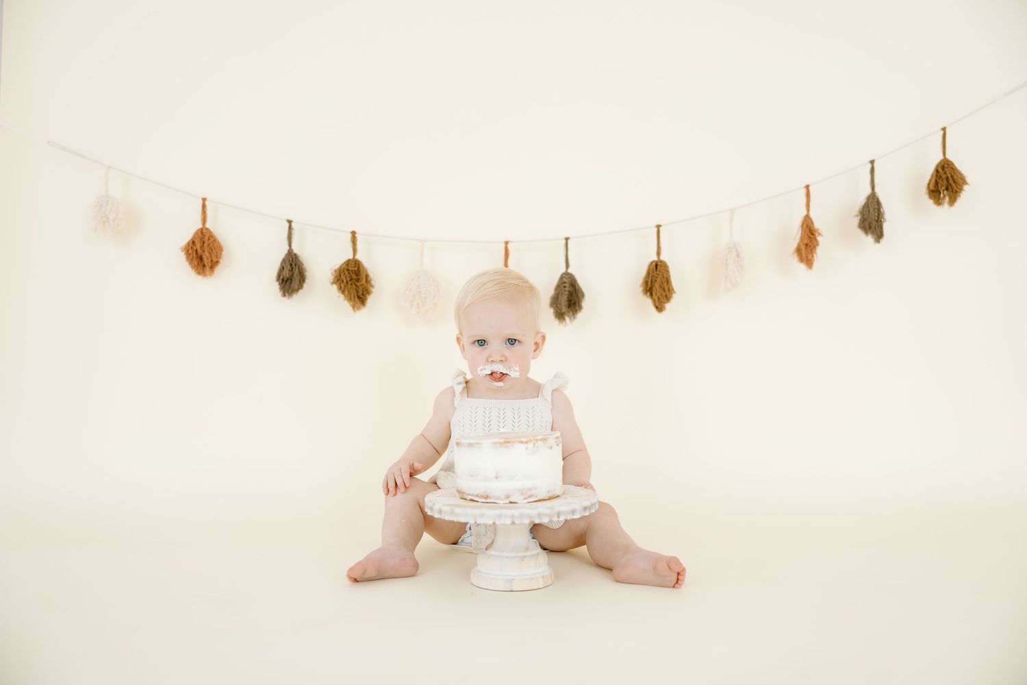 A baby in white sits on a wooden floor with autumn leaf garland decorations hanging overhead.